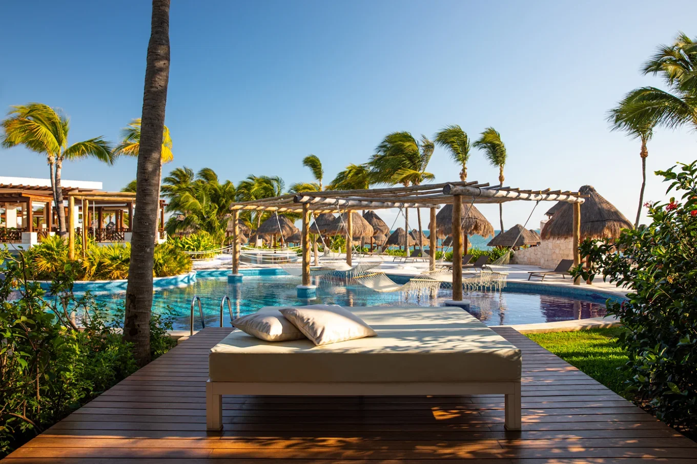 Poolside lounge chair with pillows at a tropical resort featuring palm trees, thatched umbrellas, and a swimming pool in a sunny setting.