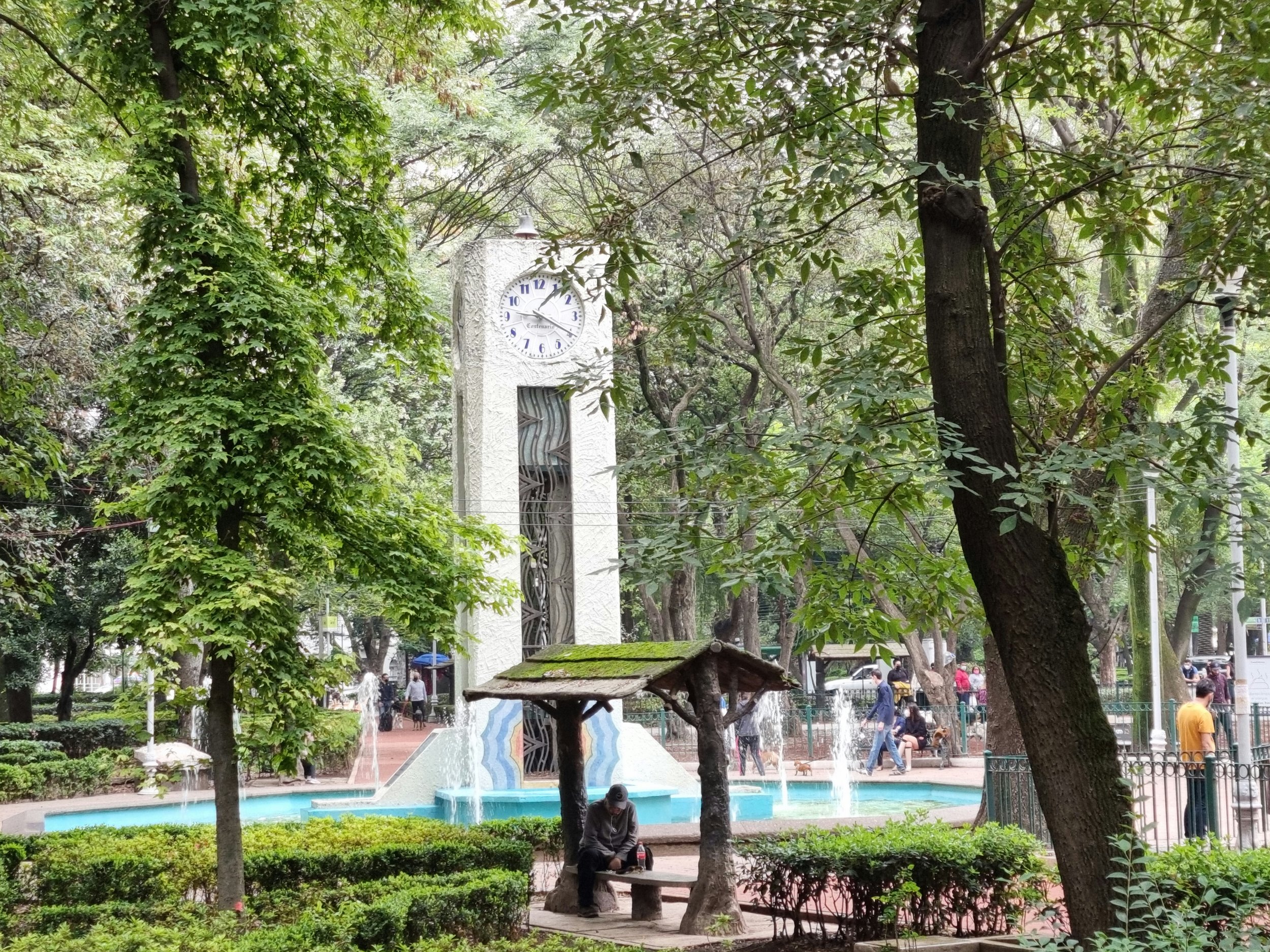 A park with a clock tower in the background, trees and greenery in the foreground, a fountain, a small bench, and people walking and sitting.