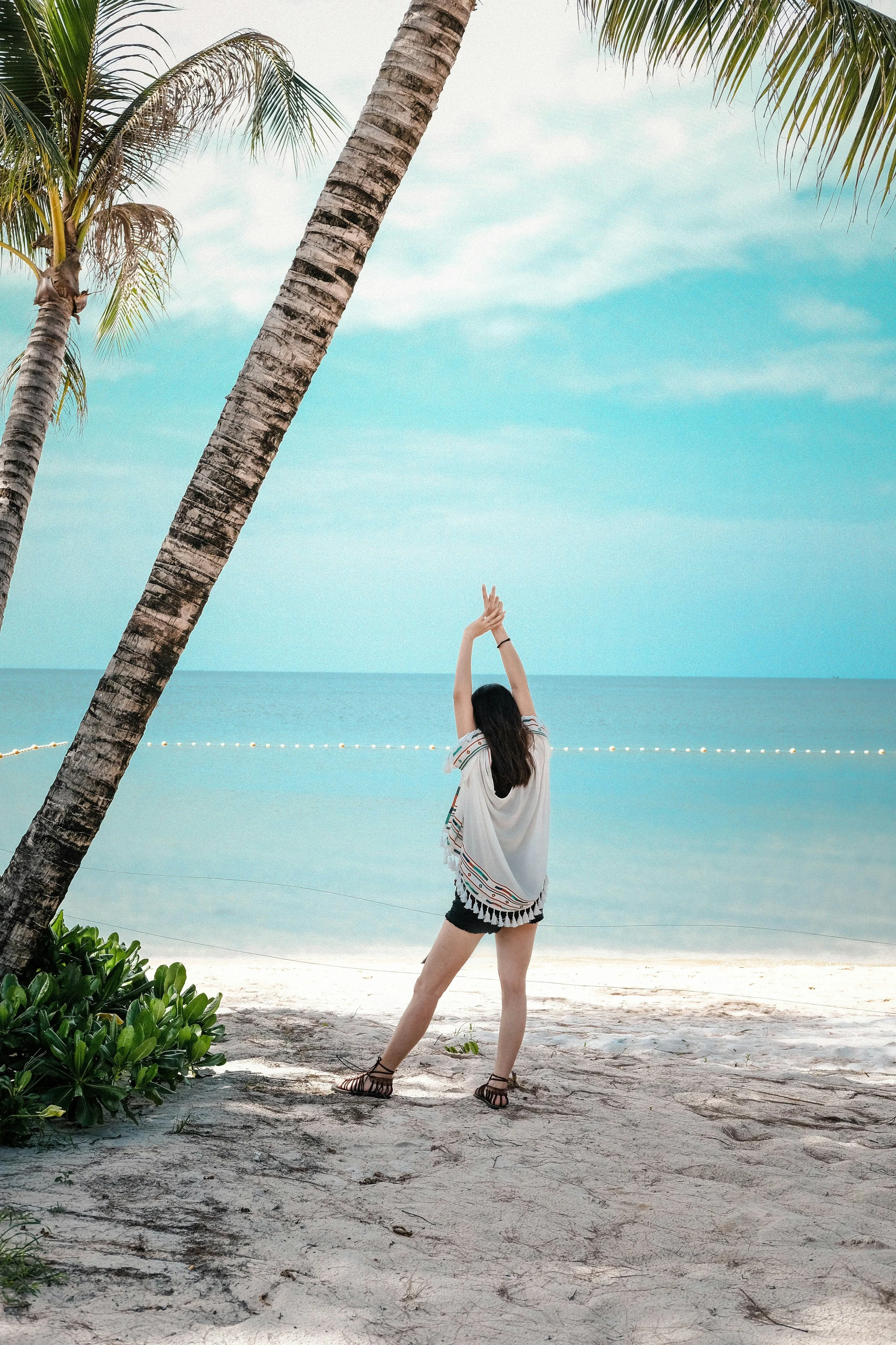 A woman stretching or dancing on a sandy beach near the ocean, with palm trees and clear blue water in the background.