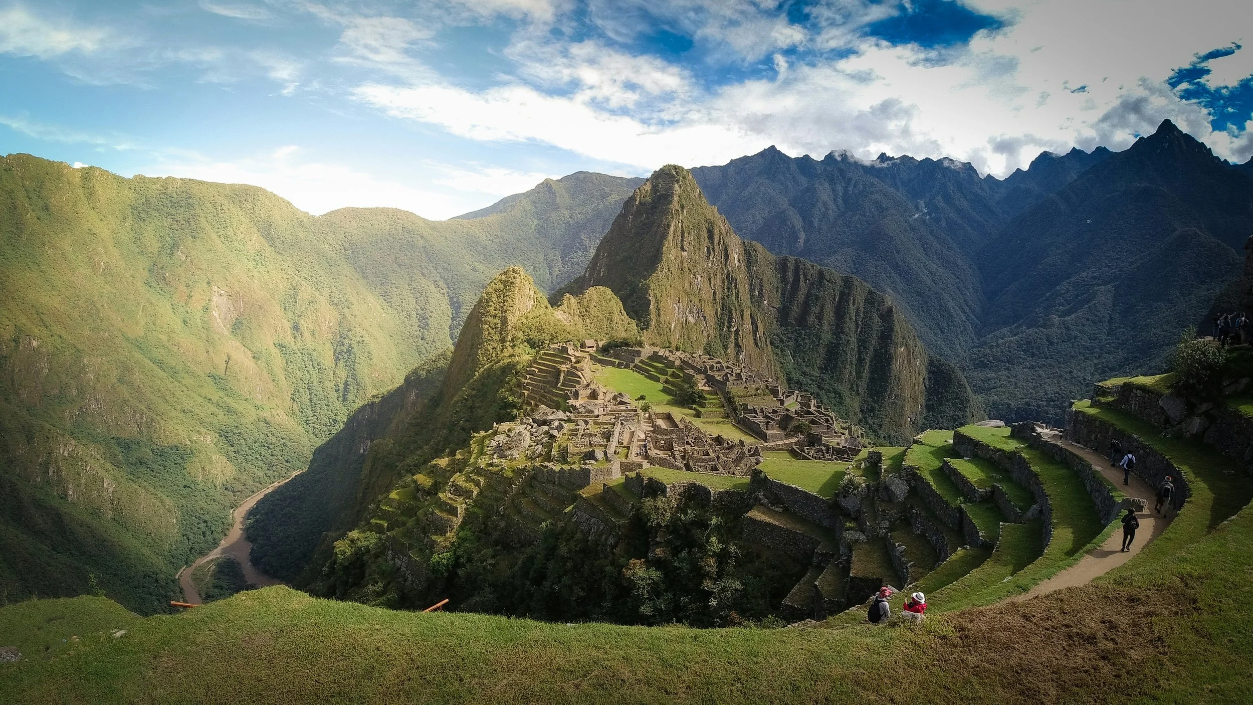 Ancient Incan ruins at Machu Picchu situated on lush green mountains with puffy clouds and a winding river below, people walking along paths exploring the site.
