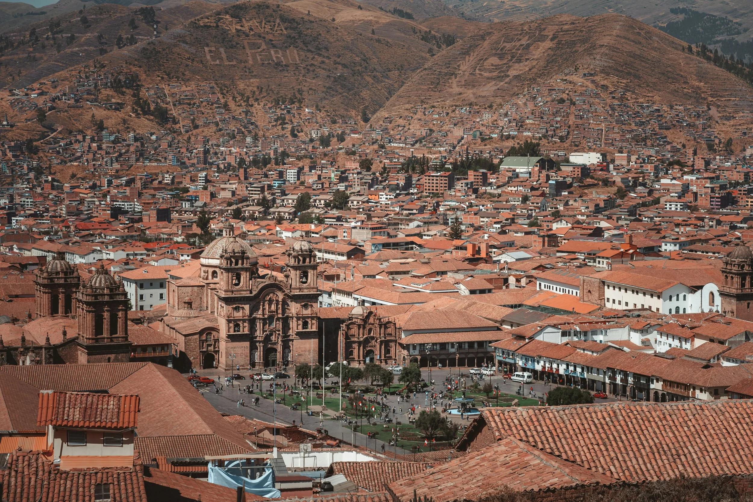 Aerial view of a city with red-tiled roofs, colonial church buildings, and a large square with people and trees, surrounded by hills with dry vegetation.