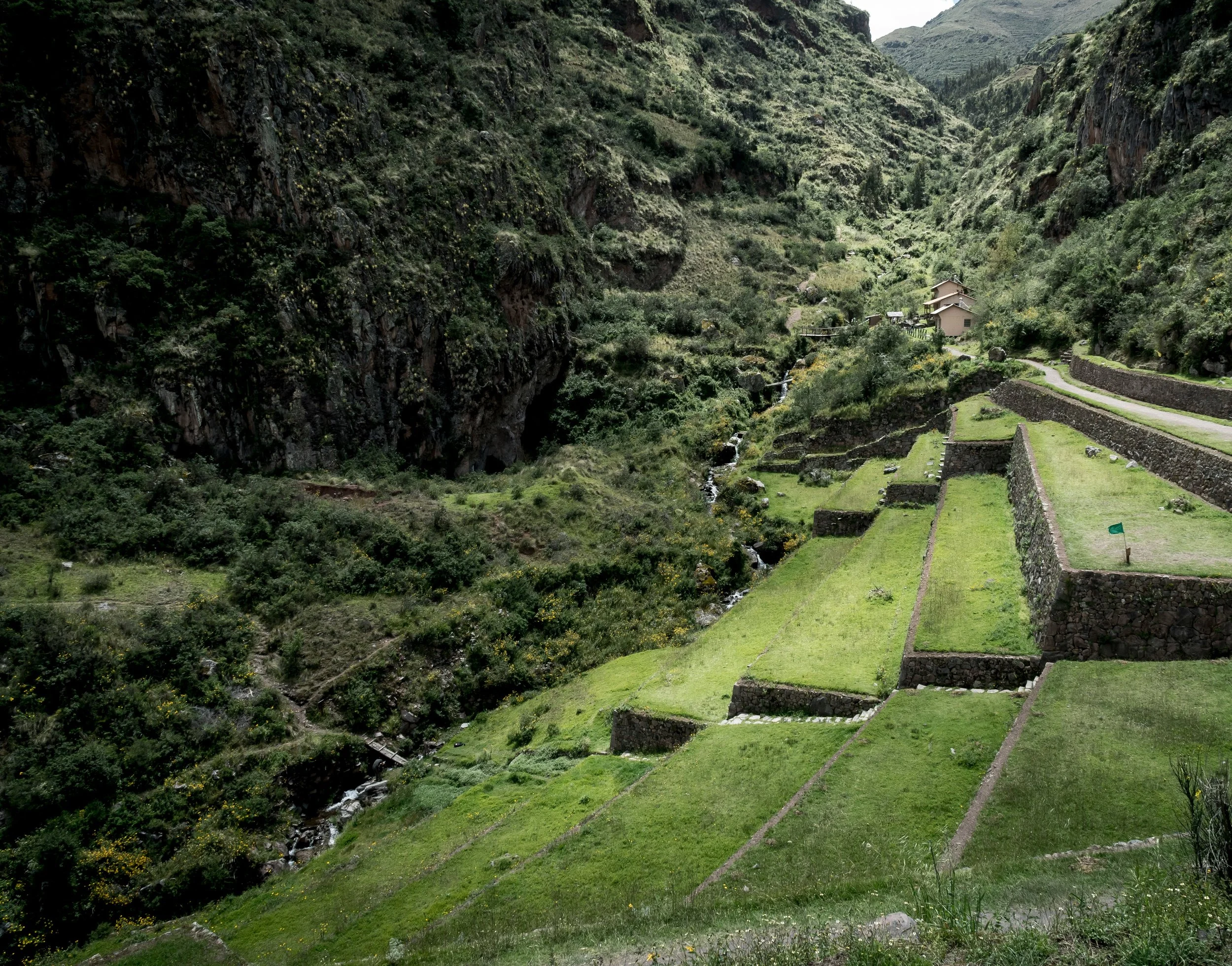 Terraced green fields and stone walls in a valley with tall mountains and a small stream.