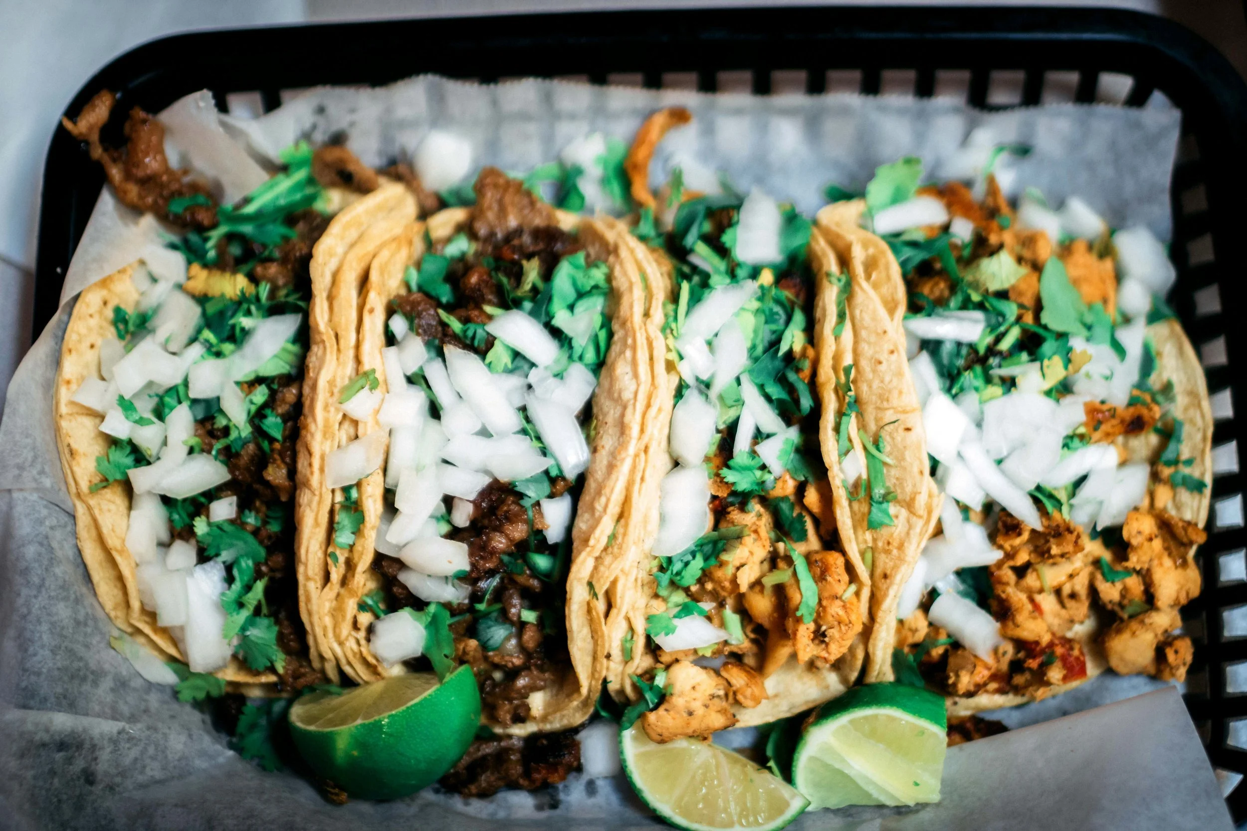Four street-style tacos in a basket topped with chopped onions and cilantro, with lime wedges on the side.