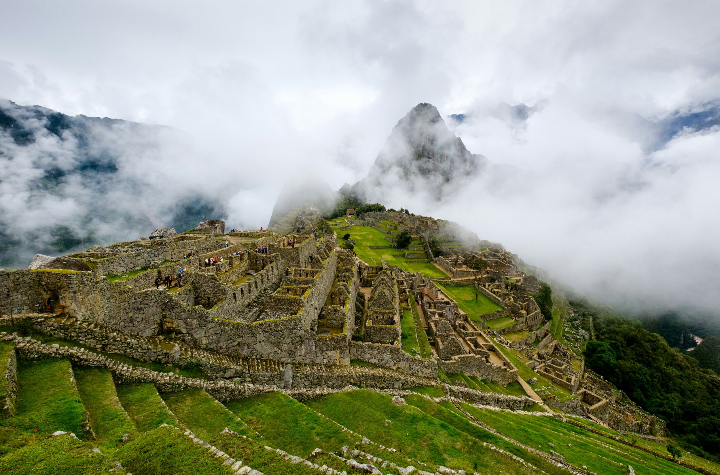 Ancient stone ruins of Machu Picchu with terraced fields and steep mountain in the background, partially obscured by clouds.