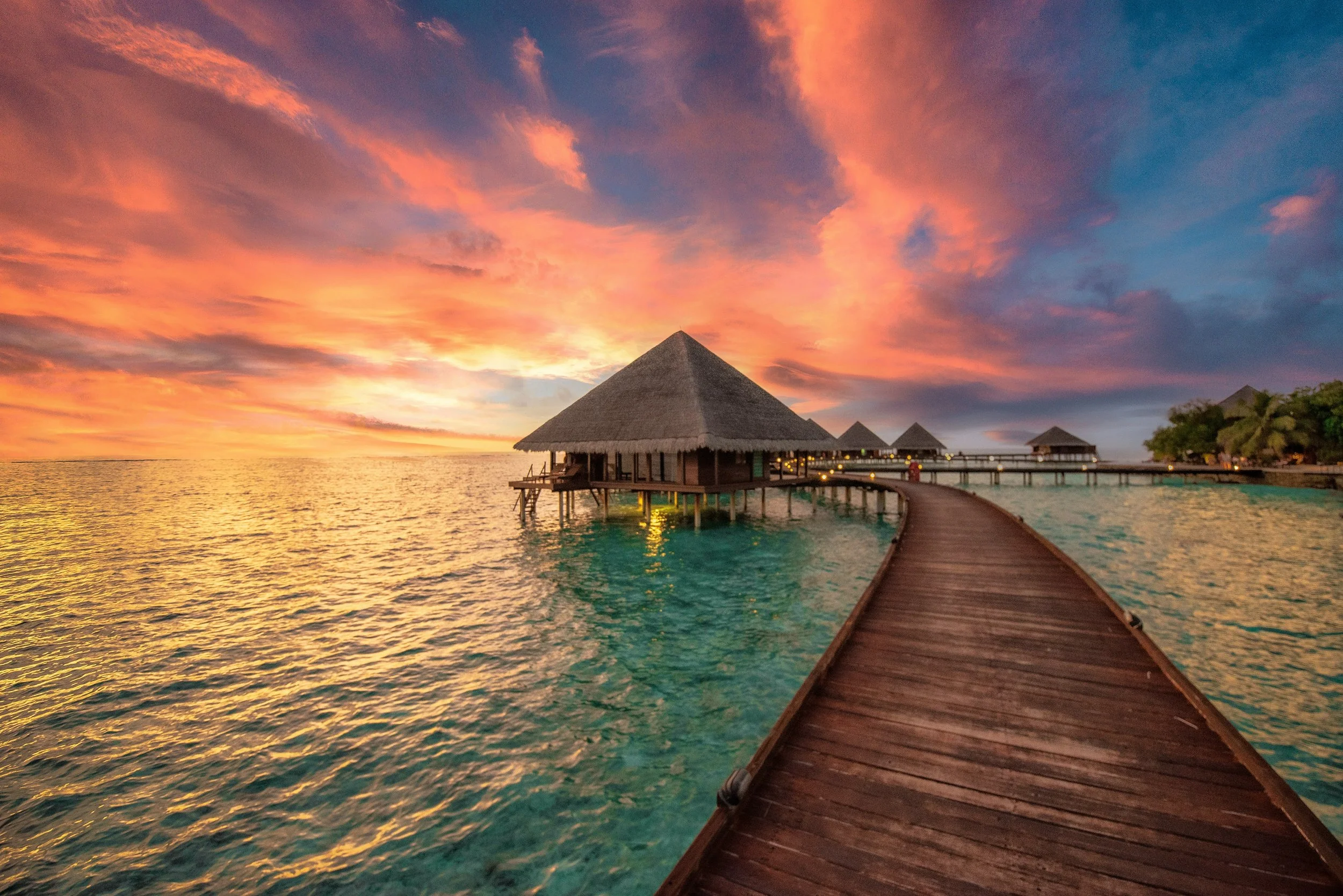 Overwater bungalows at sunset with a wooden walkway leading to the structures, the sky filled with colorful clouds, and calm ocean waters below.