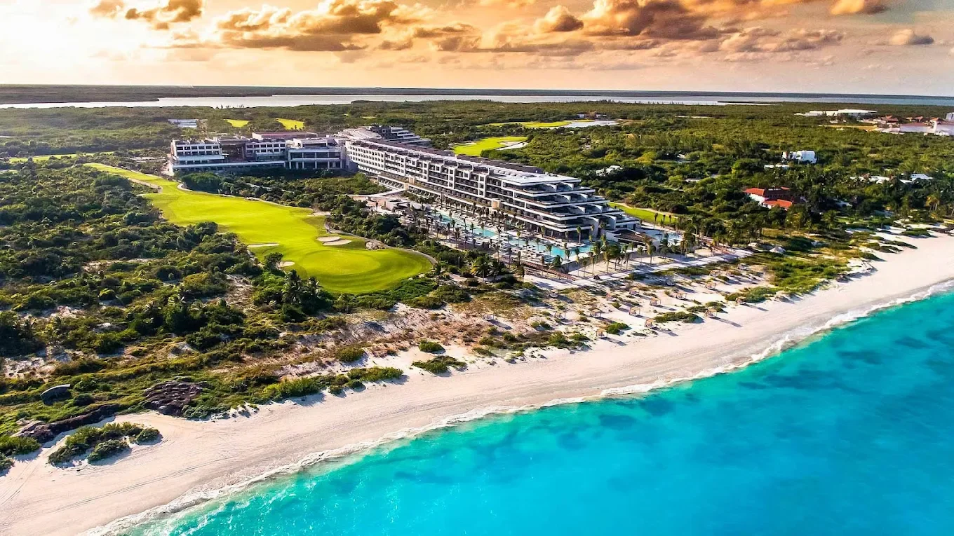 Aerial view of a modern beachfront hotel with a golf course, sandy beach, and turquoise ocean water at sunset.