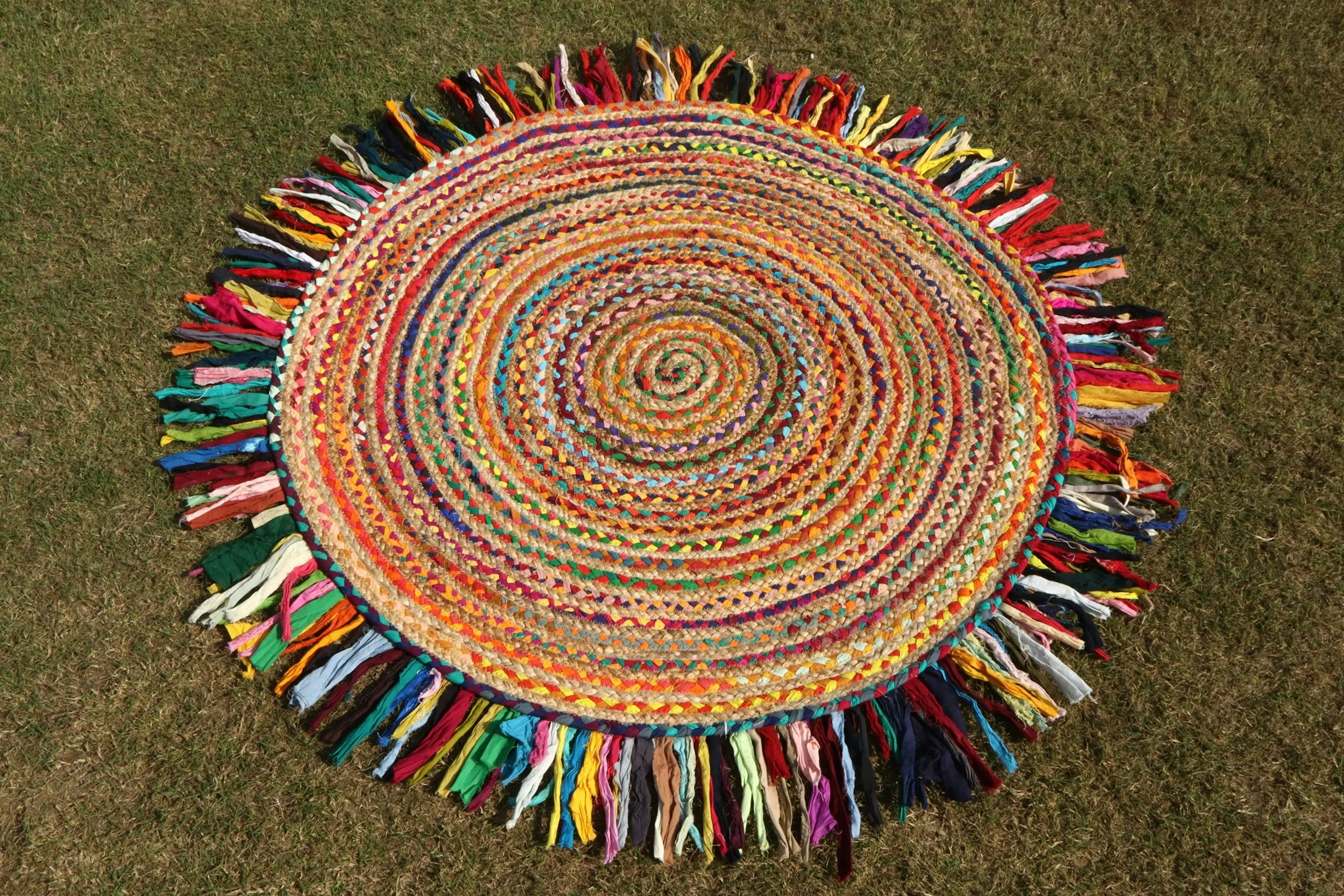 Colorful round braided rug with multicolored fabric fringe edges, placed on grass.