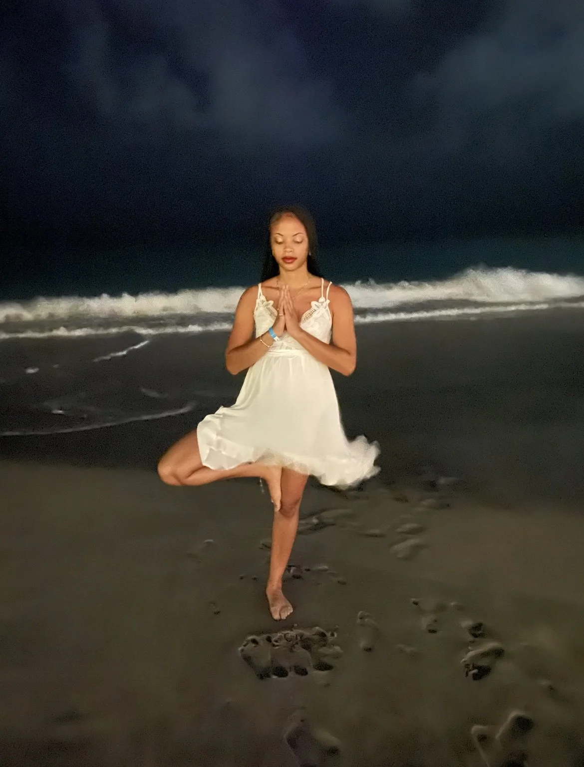 A woman in a white dress practicing yoga on a beach at night with dark sky and waves in the background.