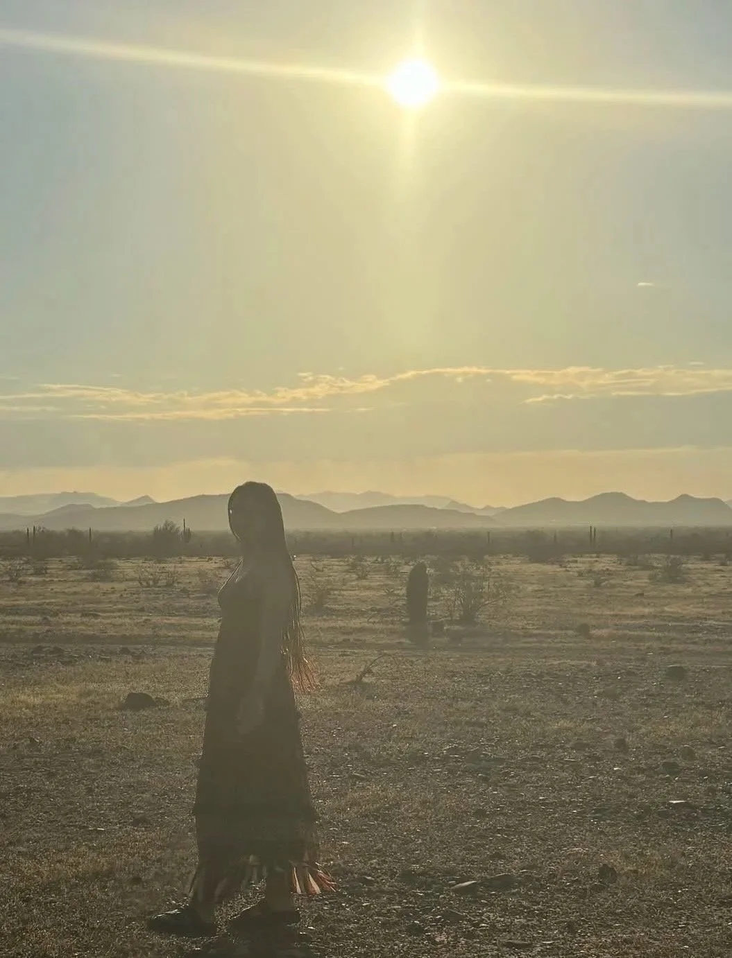 Woman standing in a desert landscape during sunset with mountains in the background.