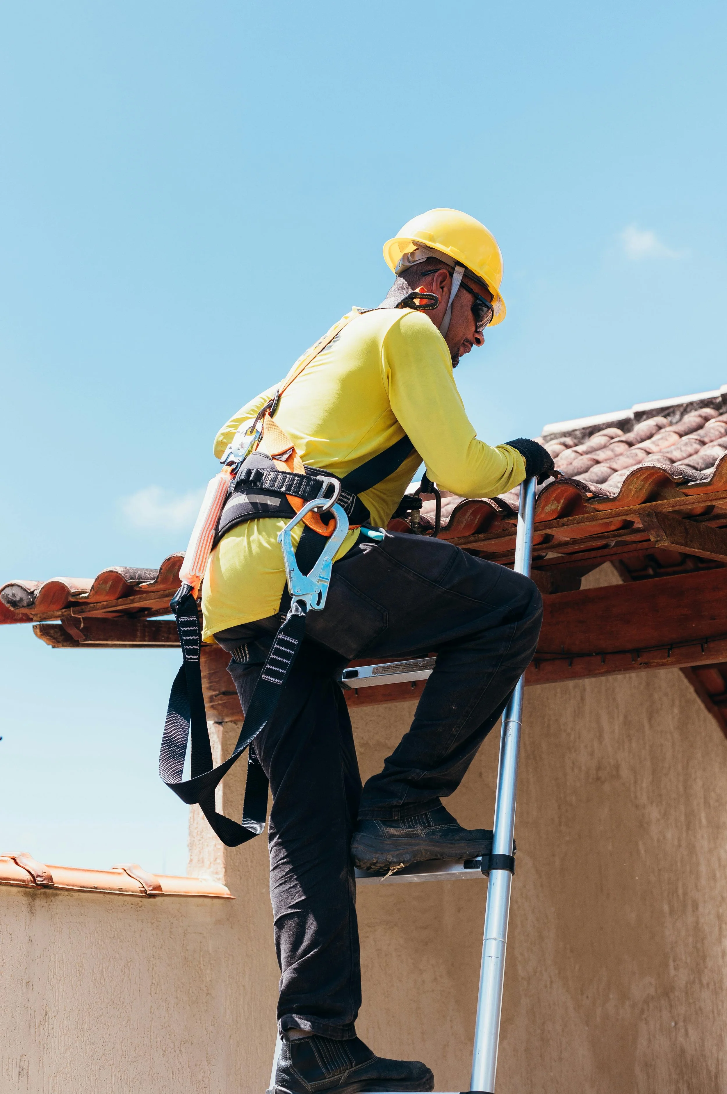 A construction worker in a yellow hard hat and safety gear installing or repairing a roof on a house under a blue sky.