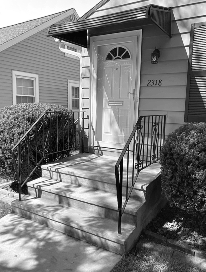 Front entrance of a house with concrete steps, metal railings, a white door with a semi-circular window, shrubs on both sides, house number 2318, and a wall-mounted light fixture.