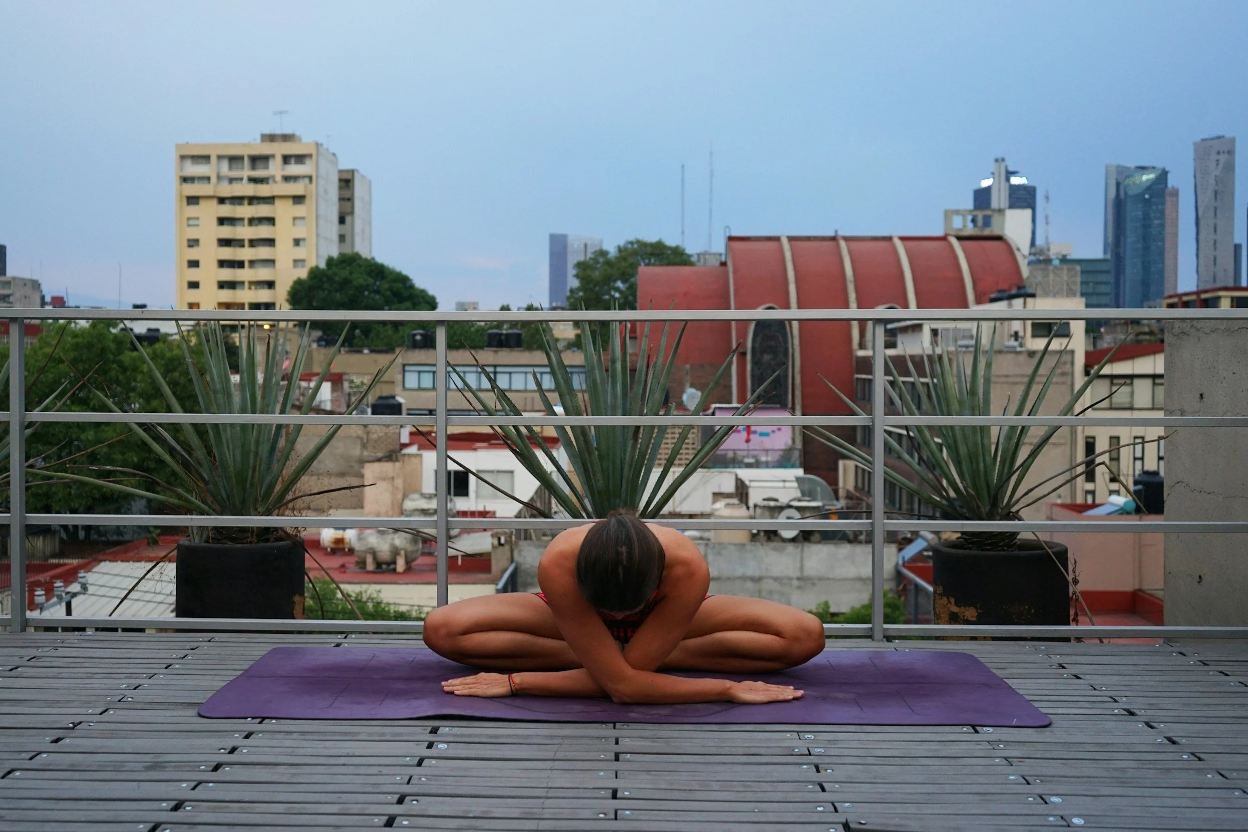 A woman practicing yoga in a seated meditation pose on a yoga mat, sitting cross-legged with hands resting on her knees, in a room with wooden floor and plain wall.
