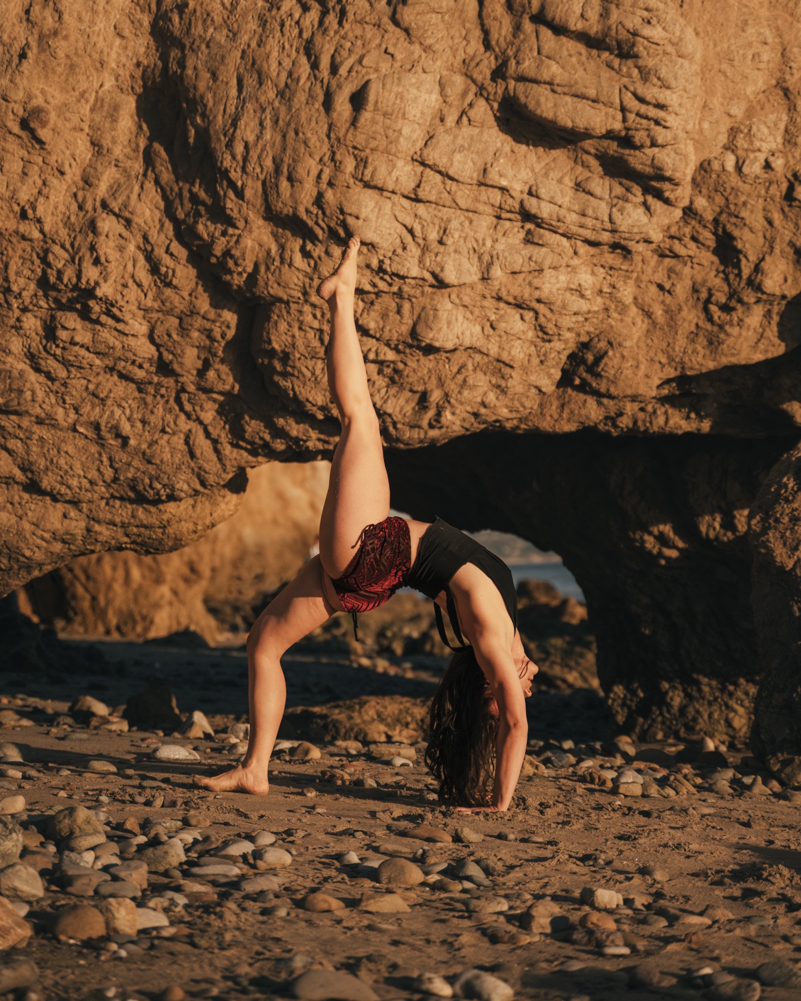 Woman practicing yoga in a downward dog position beneath large rock formations on a sandy beach.