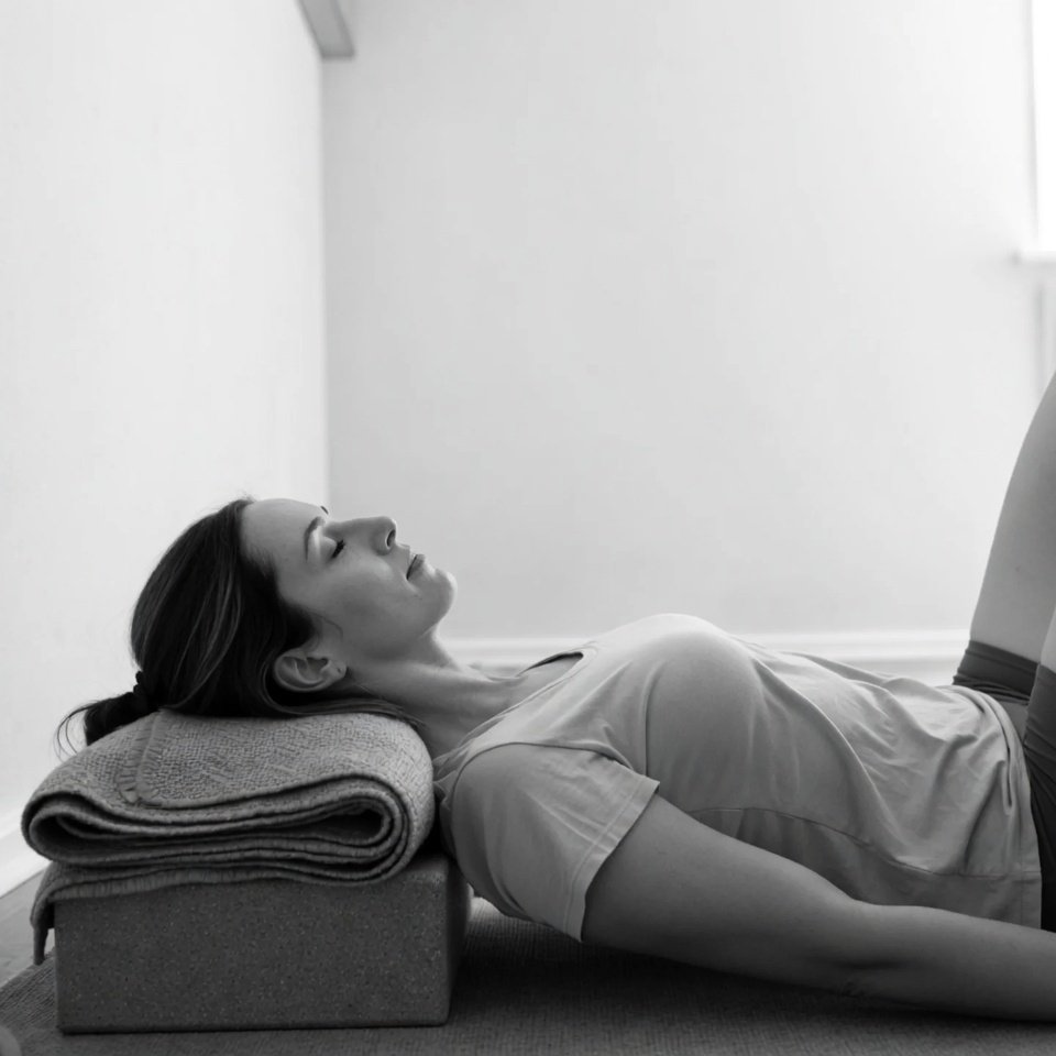 A woman lying on her back on the floor with her head resting on a rolled-up towel placed on a cushion, in a minimalistic room.