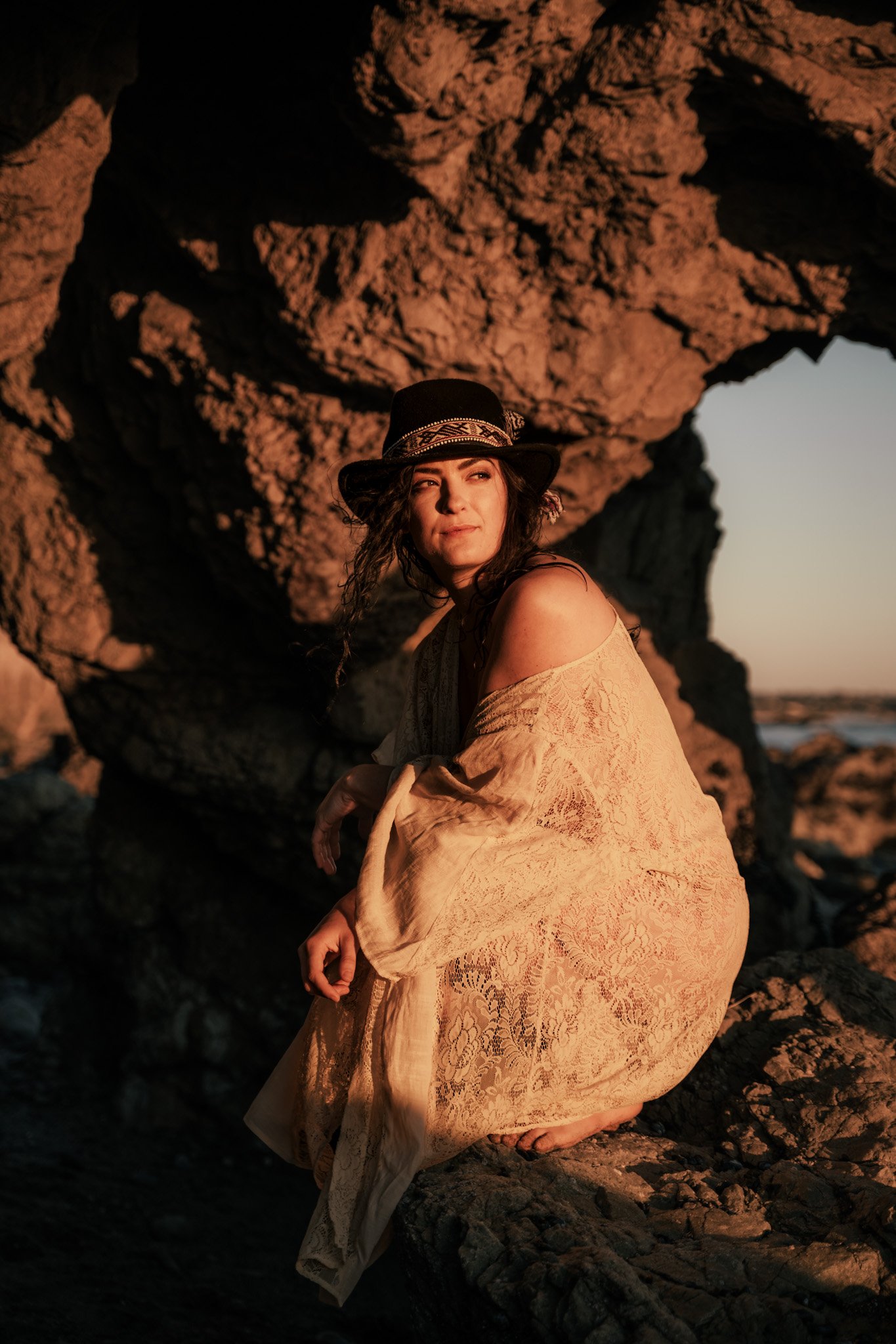 A woman sitting on rocks near the ocean during sunset, wearing a lace dress and a hat, with a rocky formation and ocean in the background.