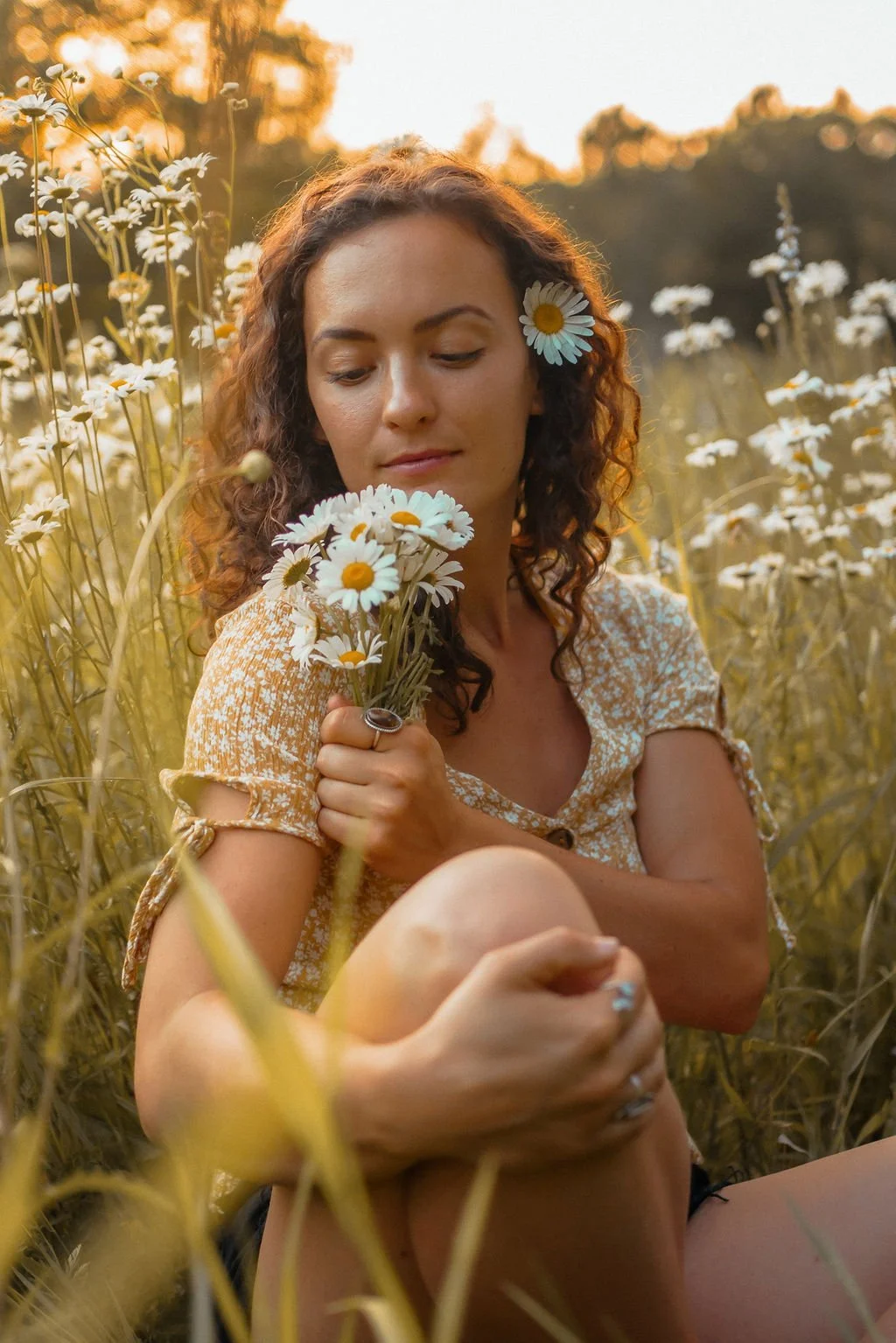 A woman with curly hair and a daisy in her hair, sitting among wild daisies in a field during sunset, holding a small bouquet of daisies and looking peacefully at the flowers.