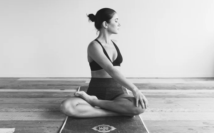 A woman practicing yoga in a seated meditation pose on a yoga mat, sitting cross-legged with hands resting on her knees, in a room with wooden floor and plain wall.