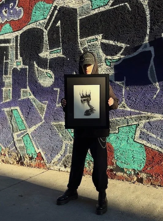 Person in dark clothing and hat holding a framed picture of a deer skull with antlers and a snake underneath, standing against a colorful graffiti wall.