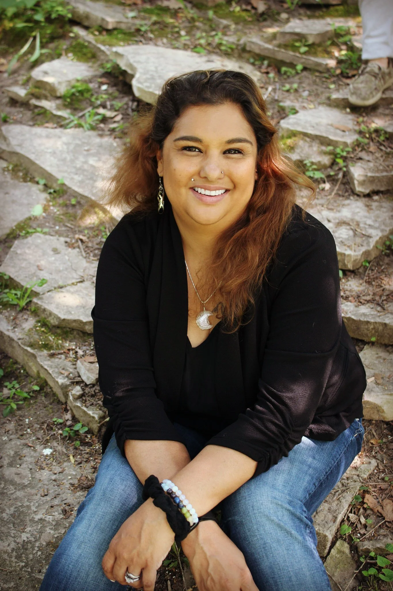 Smiling woman sitting on stone steps outdoors, wearing a black top, blue jeans, and jewelry, with greenery in the background.