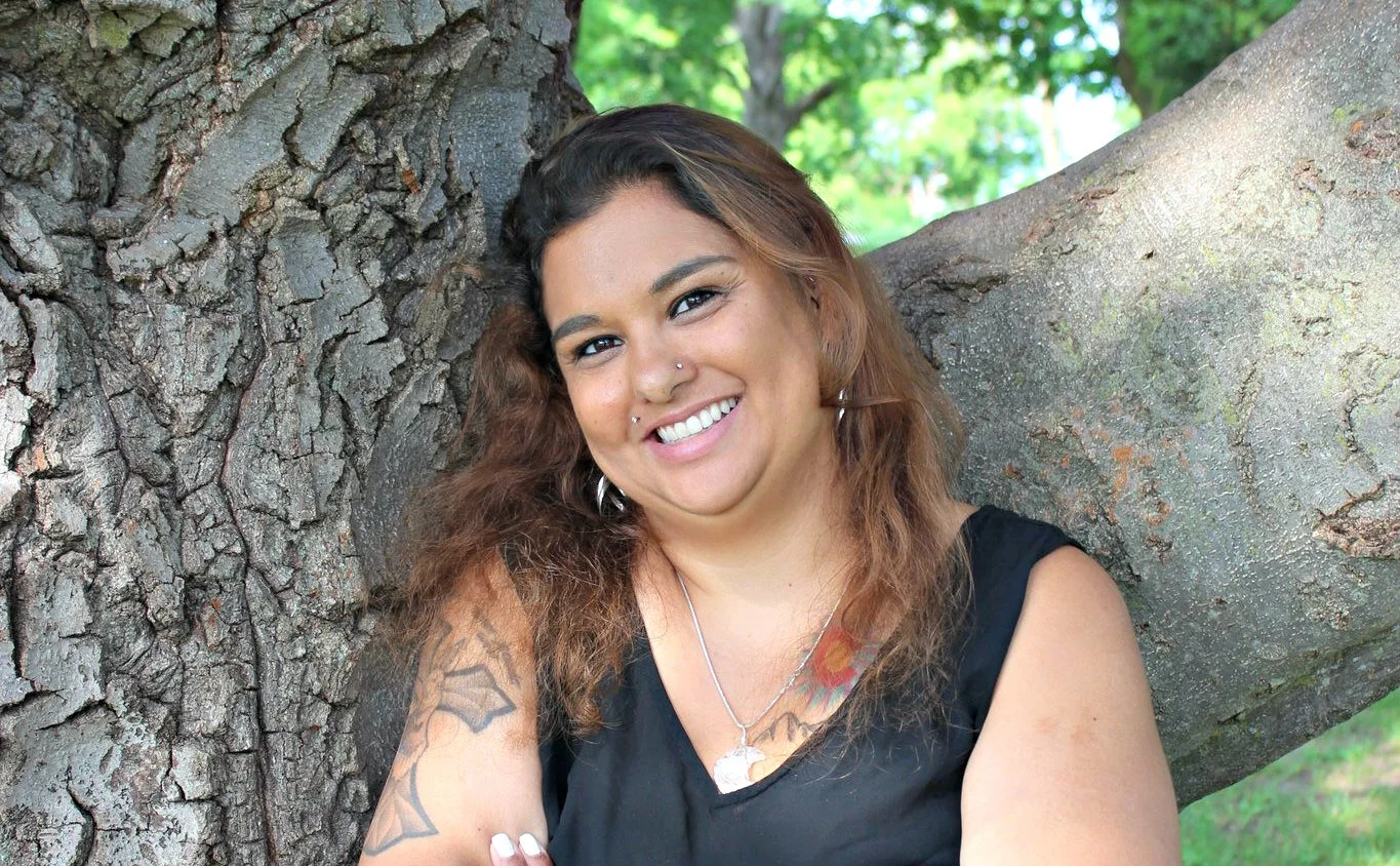 A woman with brown curly hair and a nose piercing smiling while sitting outdoors against a large tree with green foliage in the background.