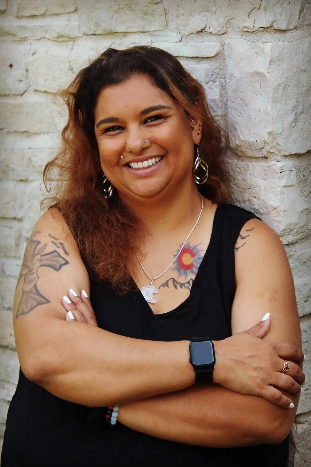 Smiling woman with multiple tattoos, wearing a black sleeveless top, jewelry, and an Apple Watch, standing against a stone wall.
