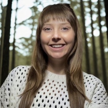 Young woman with long brown hair smiling outdoors in a forest setting, wearing a white polka dot sweater.