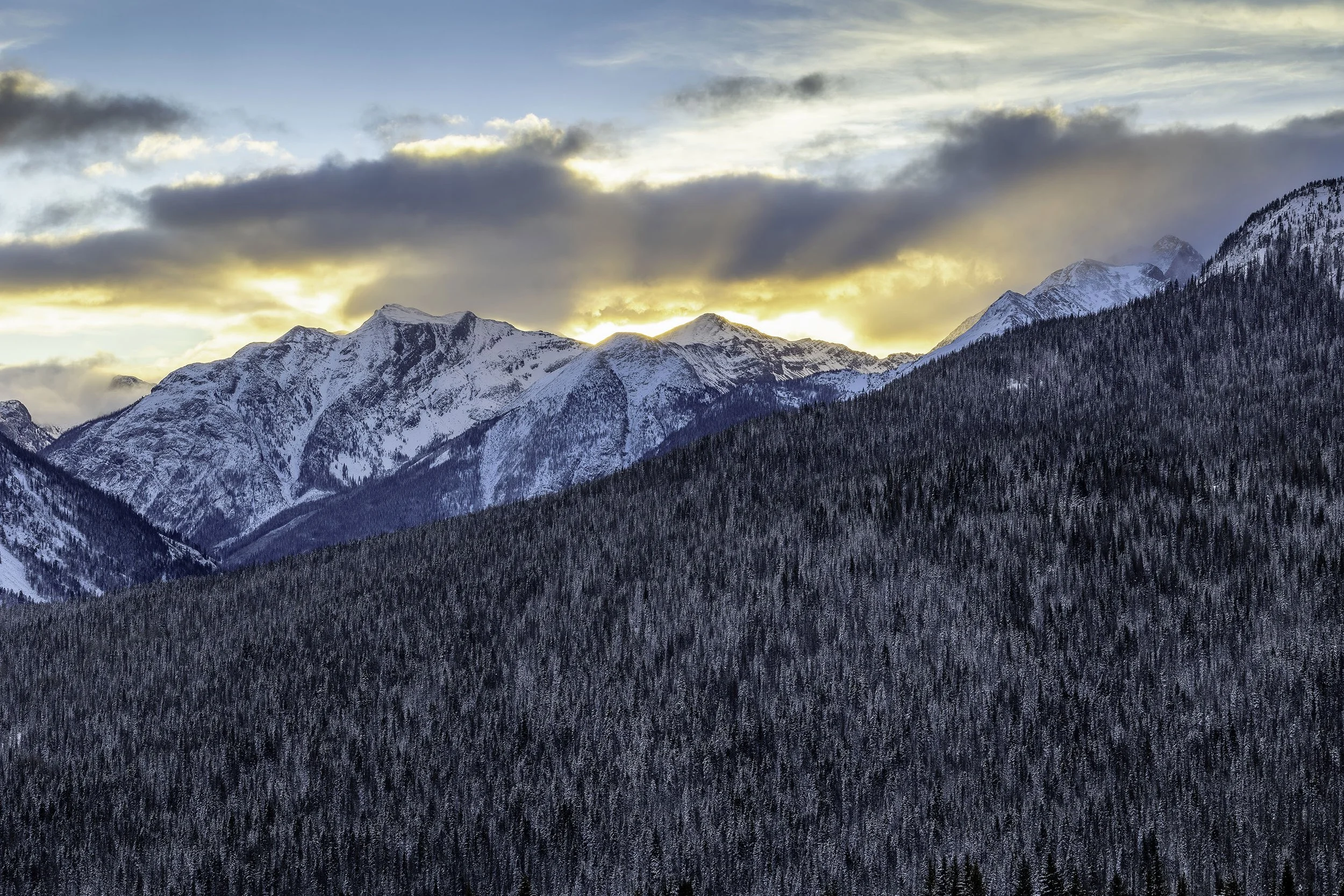 Snow-covered mountain peaks with forested slopes at sunset, partly cloudy sky