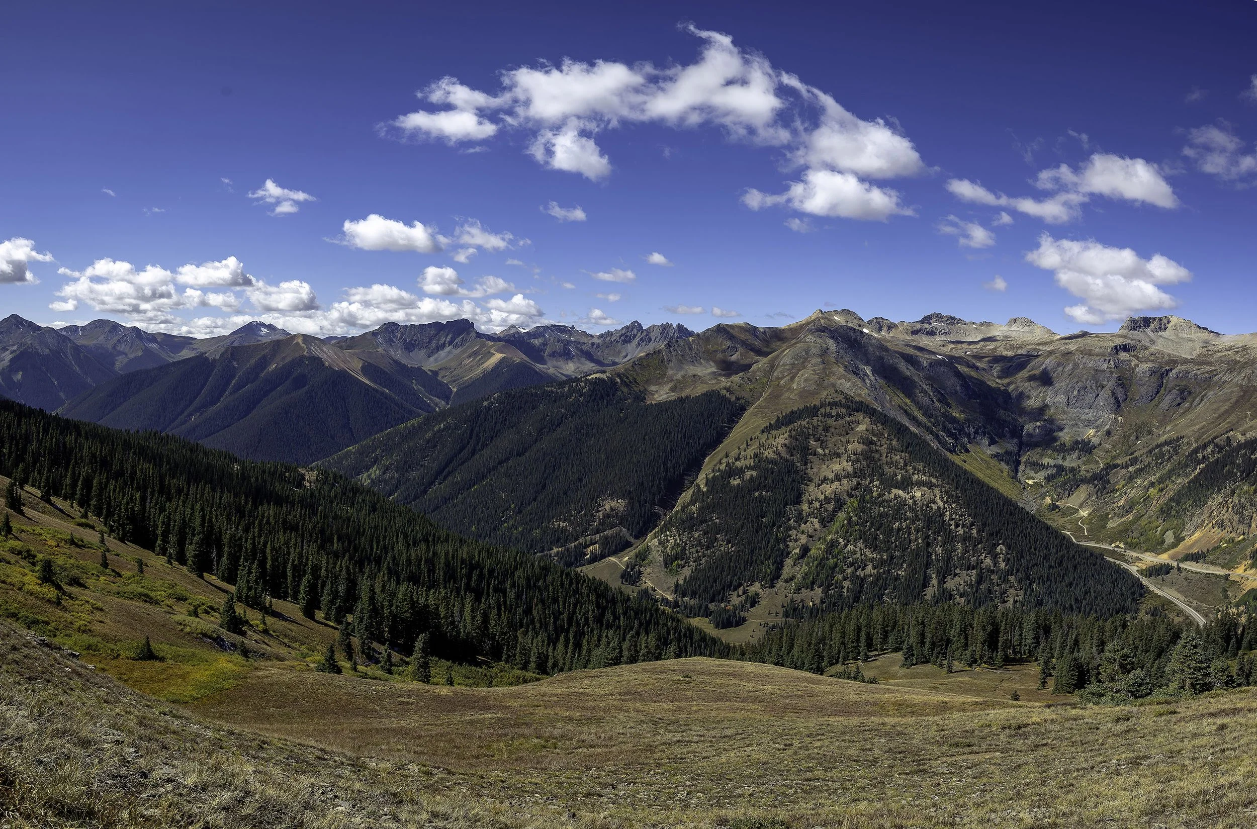 Mountain landscape with rolling hills, dense forest, and a bright blue sky with scattered clouds.