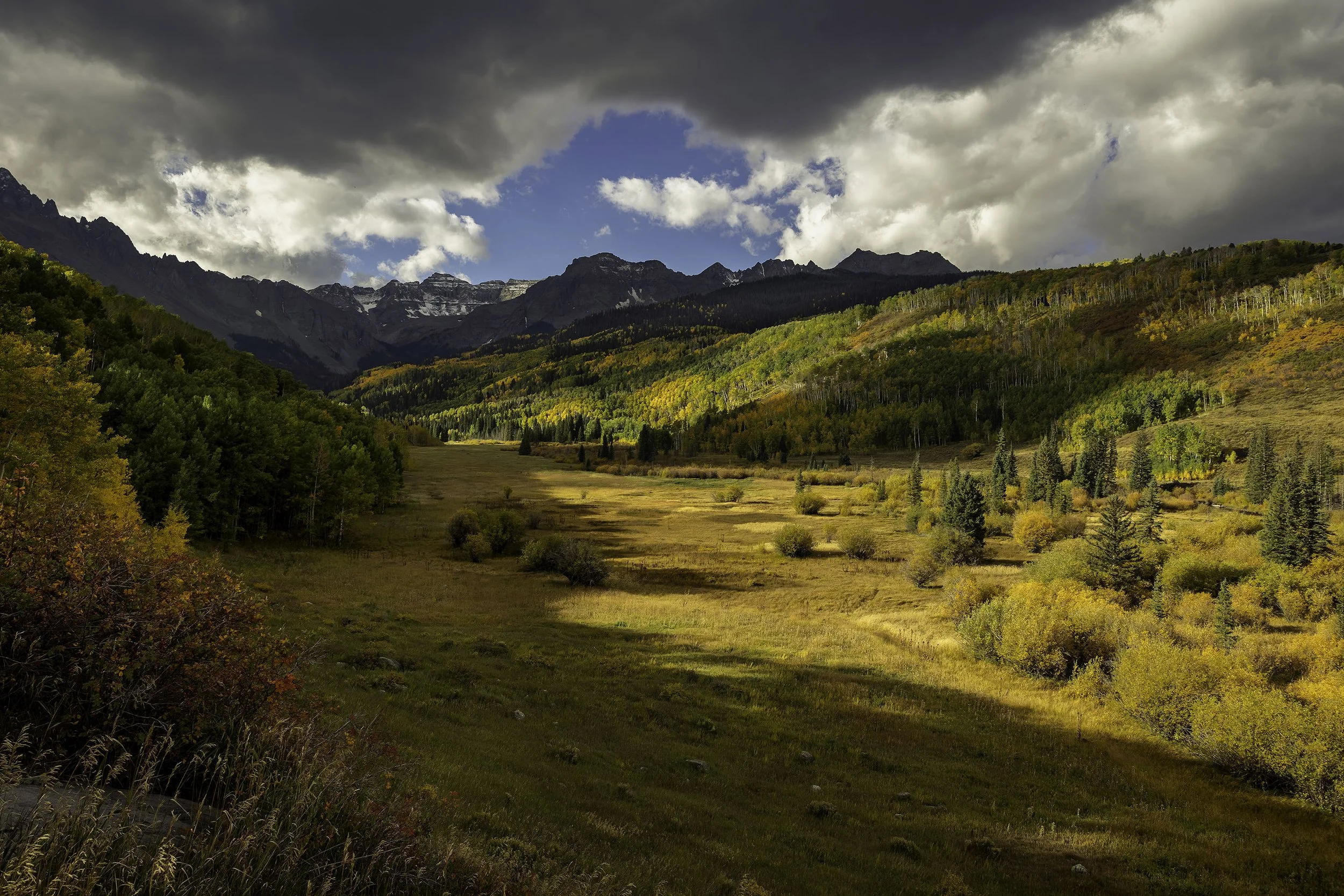 A scenic mountain landscape with a grassy valley, trees with fall foliage, and distant snow-capped peaks under dark clouds and a blue sky.