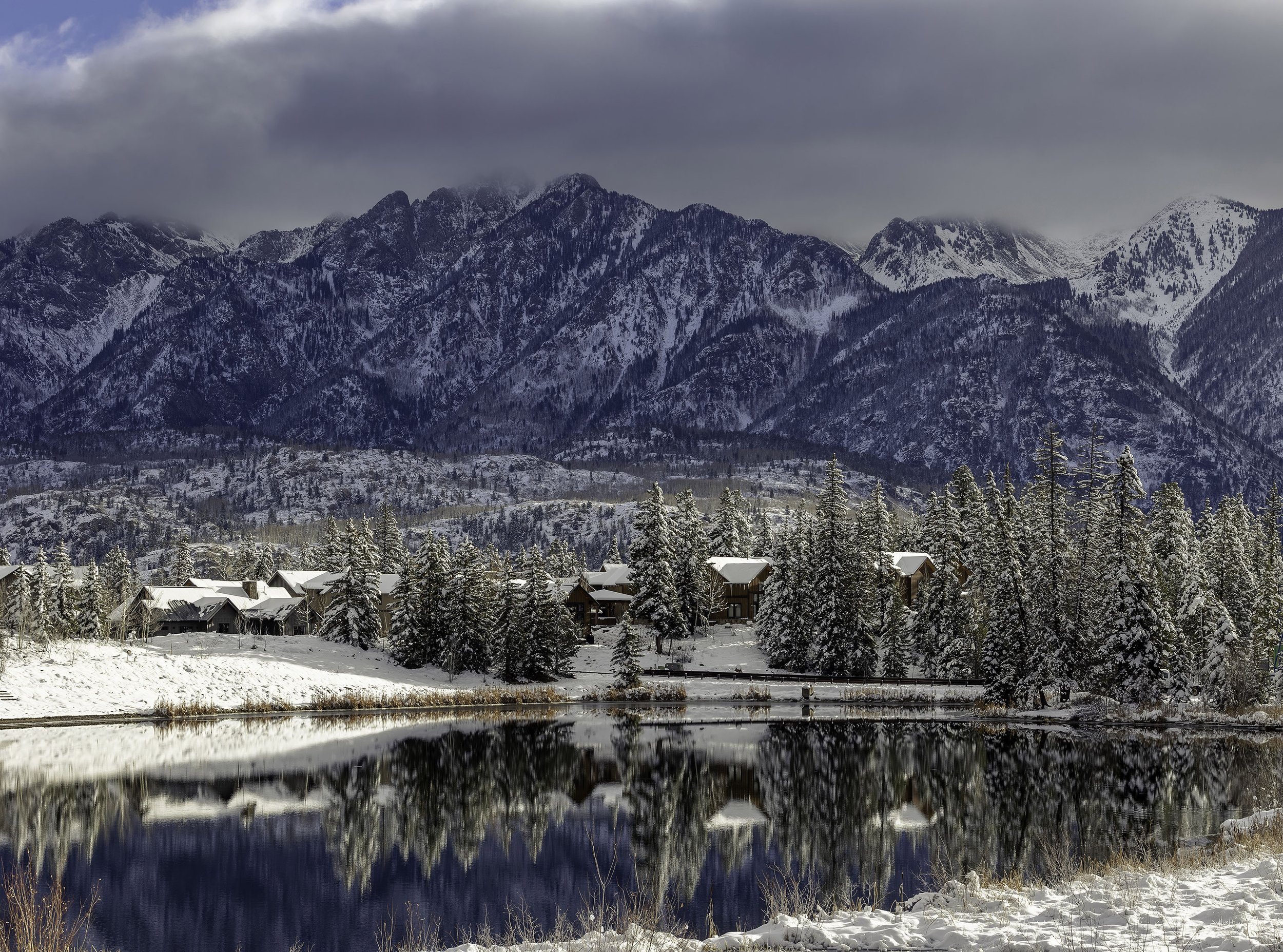 Snow-covered village with buildings and trees nestled by a calm lake, with tall snow-capped mountains in the background under a cloudy sky.