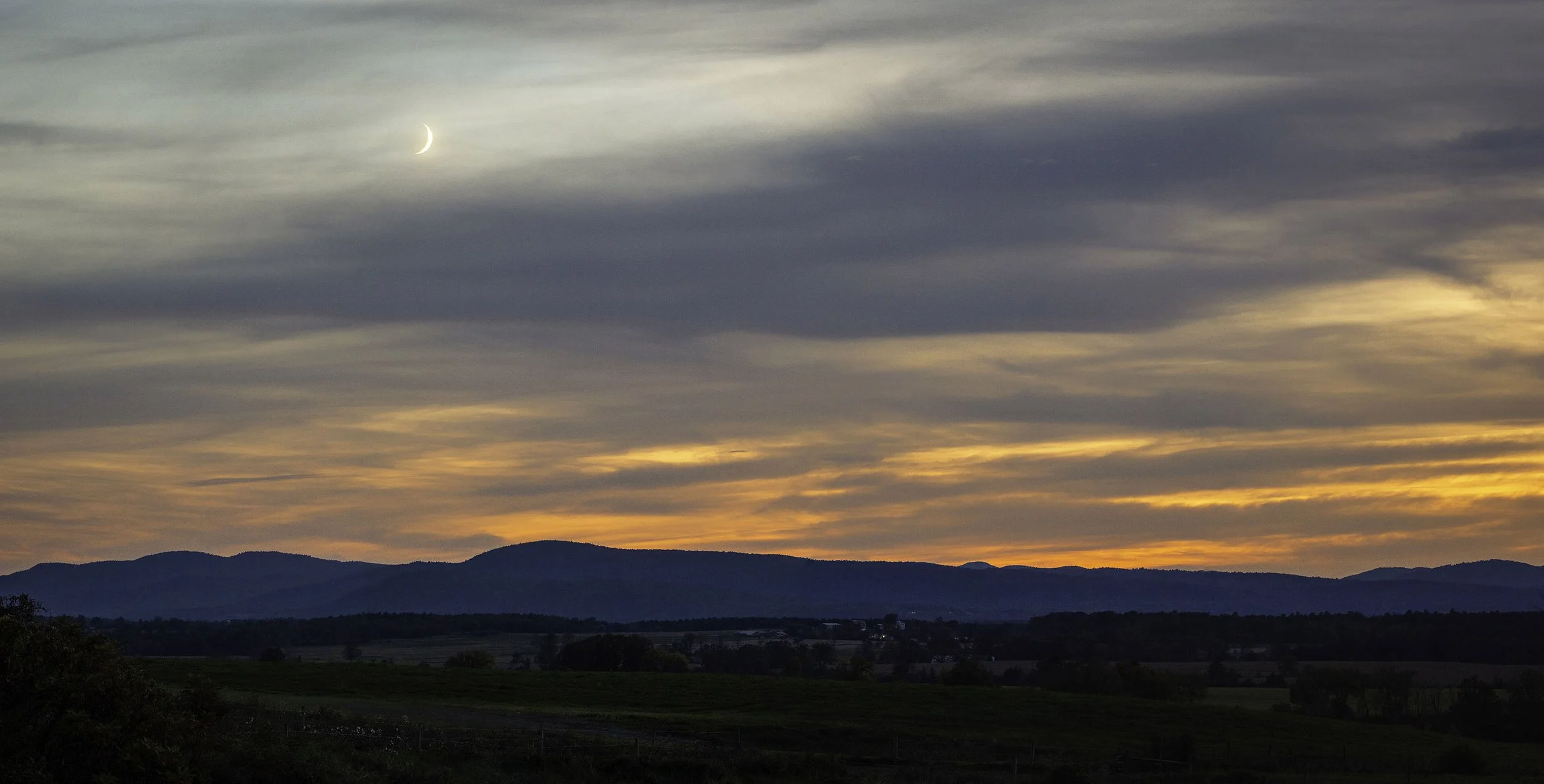 Sunset over rolling hills with a crescent moon in a partly cloudy sky.