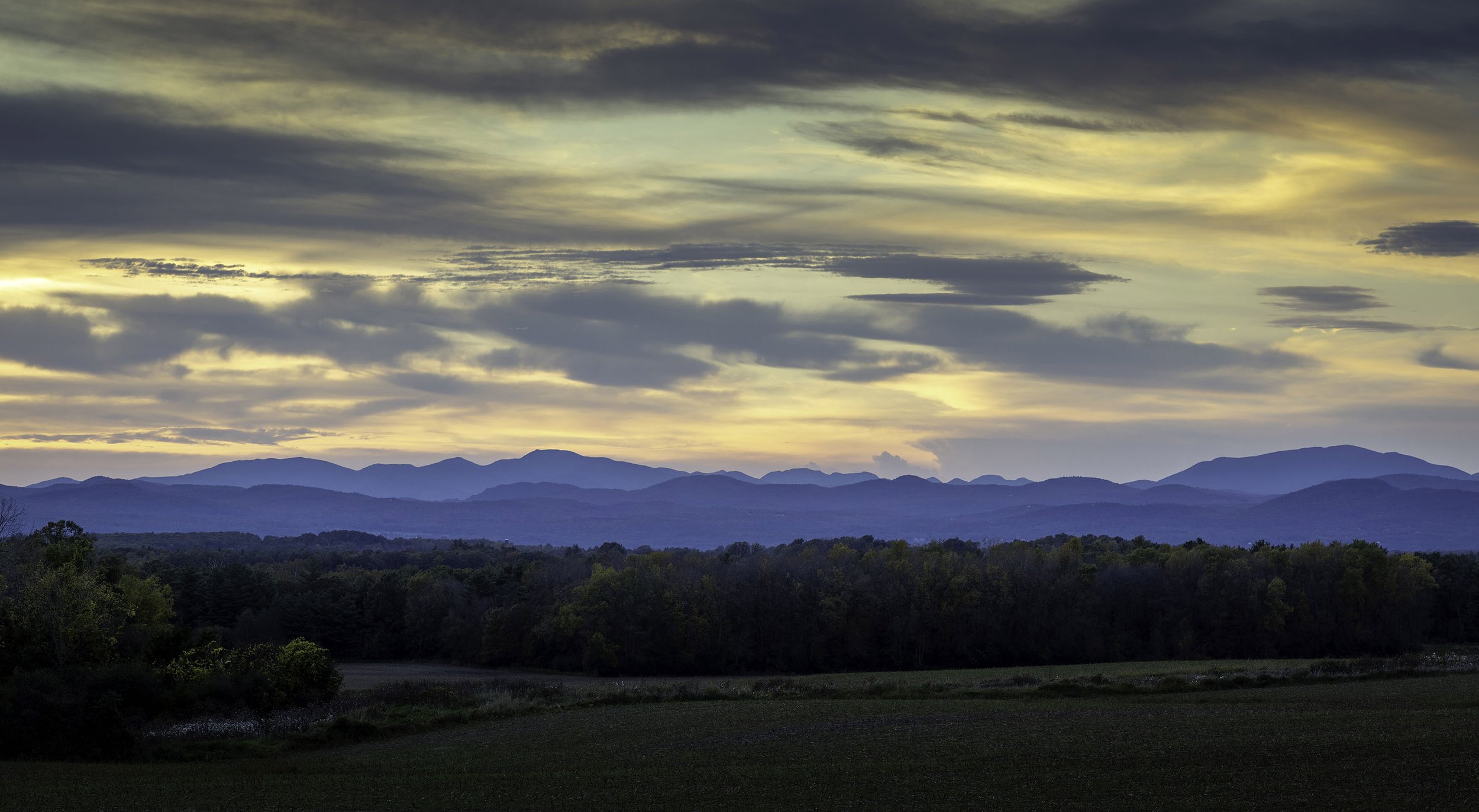 Scenic view of distant mountains under a sky with dramatic clouds during sunset, with a forested area in the foreground.