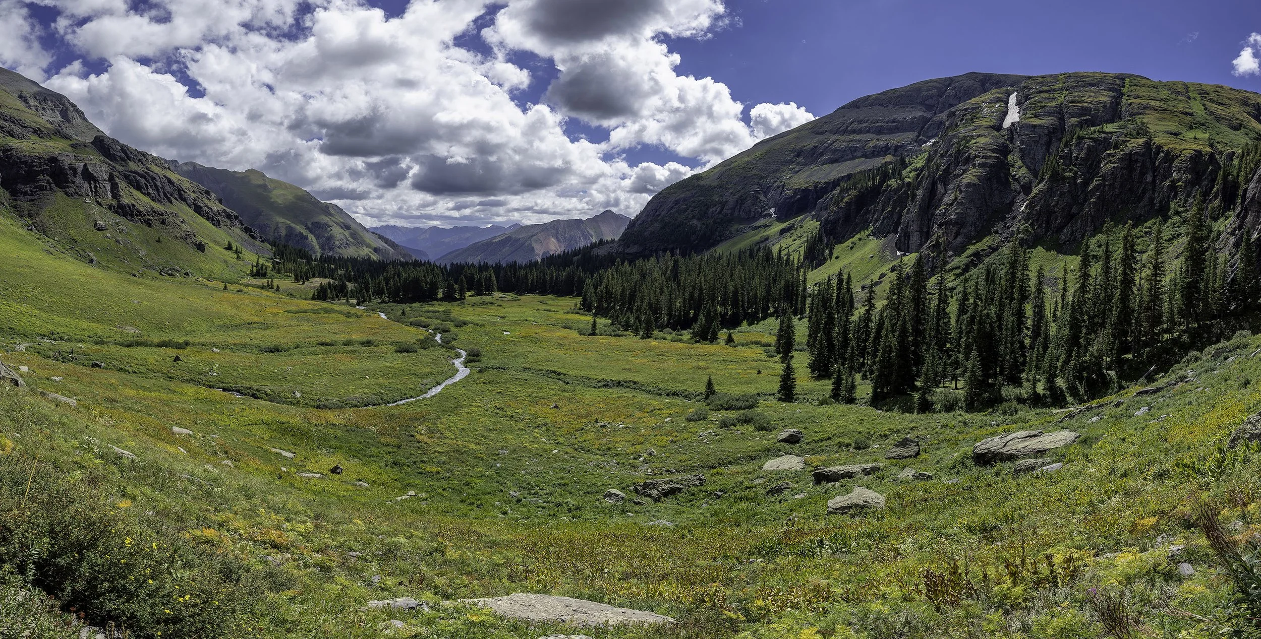 A lush valley with a meandering stream, surrounded by green mountains and forests, under a partly cloudy sky.