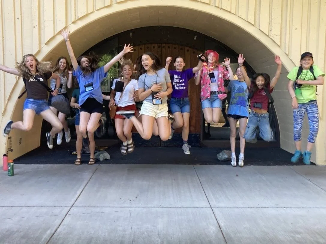 A group of children jumping in the air in front of a yellow building with a rounded entryway. They appear happy and excited.