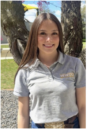 Young woman with long brown hair smiling outdoors, wearing a light gray polo shirt with a logo, standing in front of trees and a park.