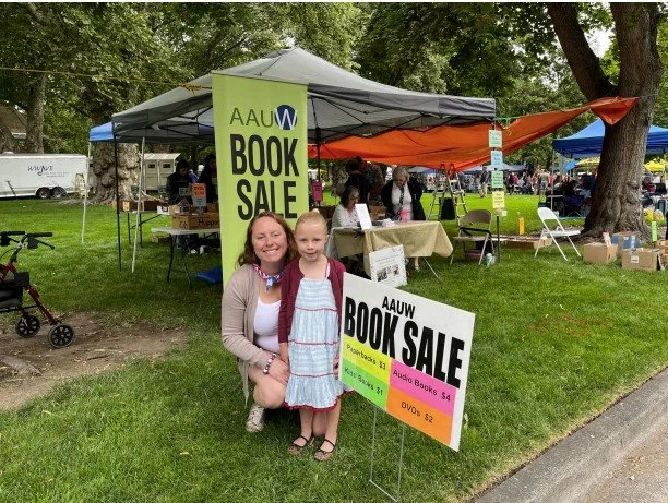 Two smiling women, one adult and one child, standing in front of a book sale tent at an outdoor event. The tent has a large yellow banner that reads 'AAUW Book Sale'. There are tables with books and people browsing in the background, along with trees and grass.