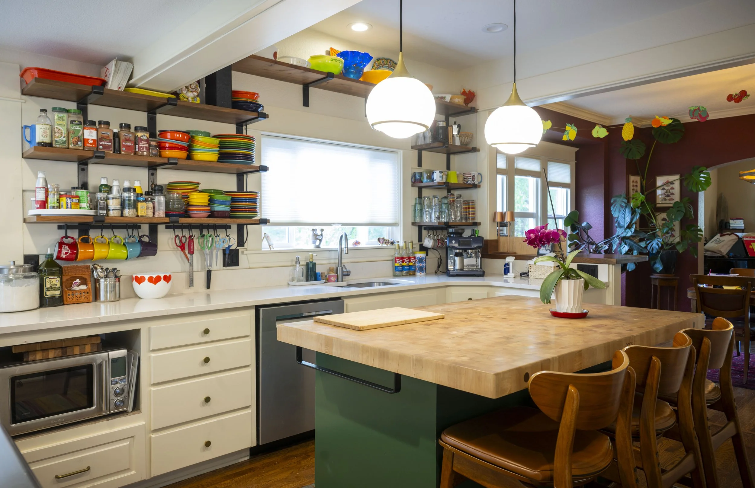 A bright kitchen with open wooden shelves holding colorful dishes, bowls, and mugs. There is a white countertop with small kitchen gadgets, a potted orchid on a wooden island, and large windows with roller shades. Pendant lights hang from the ceiling