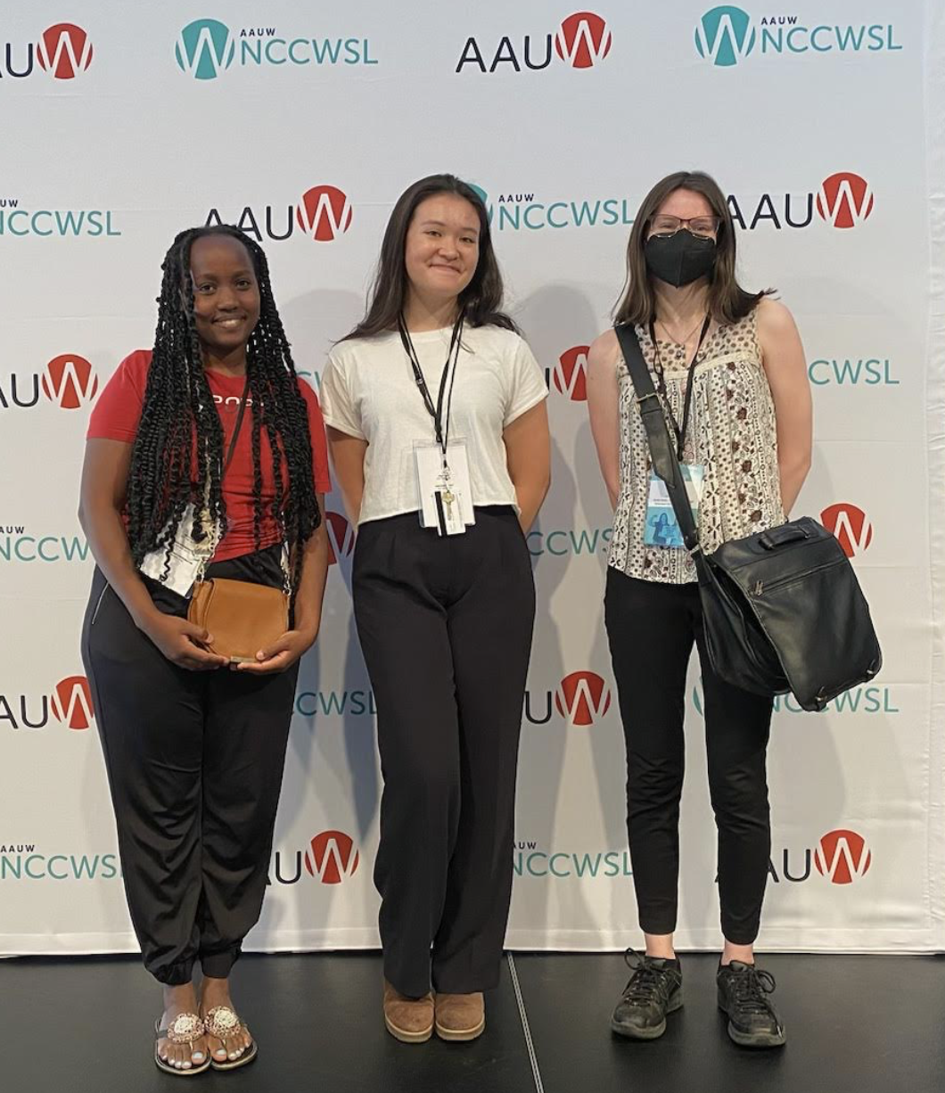 Three women standing in front of a step-and-repeat banner with logos for AAUW, NCCWSL, and AUW, at an event. They are smiling, with one wearing a mask.