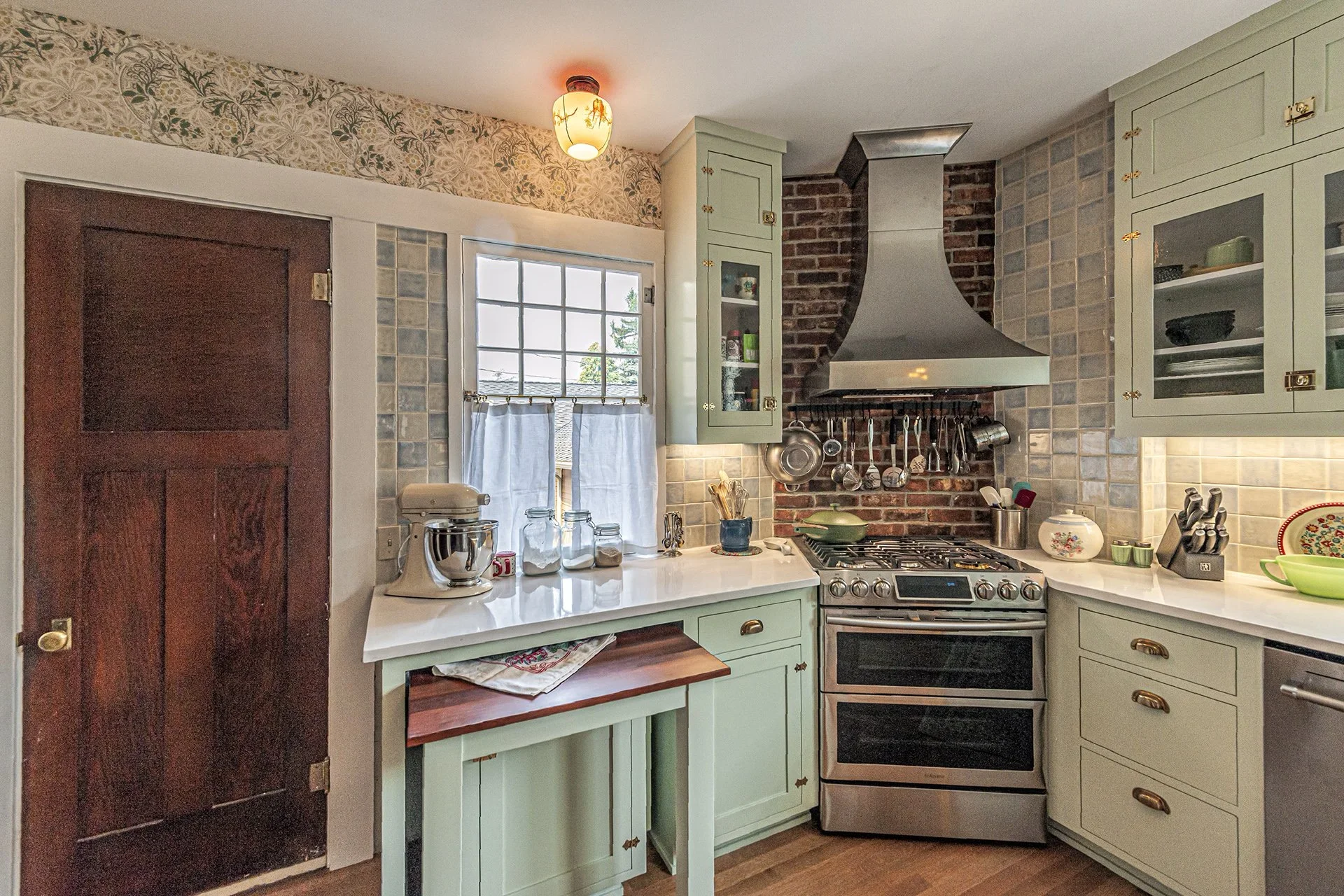 A cozy kitchen with light green cabinets, a brick and stainless steel range hood above a stove, a window with white curtains, a mix of modern and vintage decor, and a floral wallpaper border at the top of the wall.