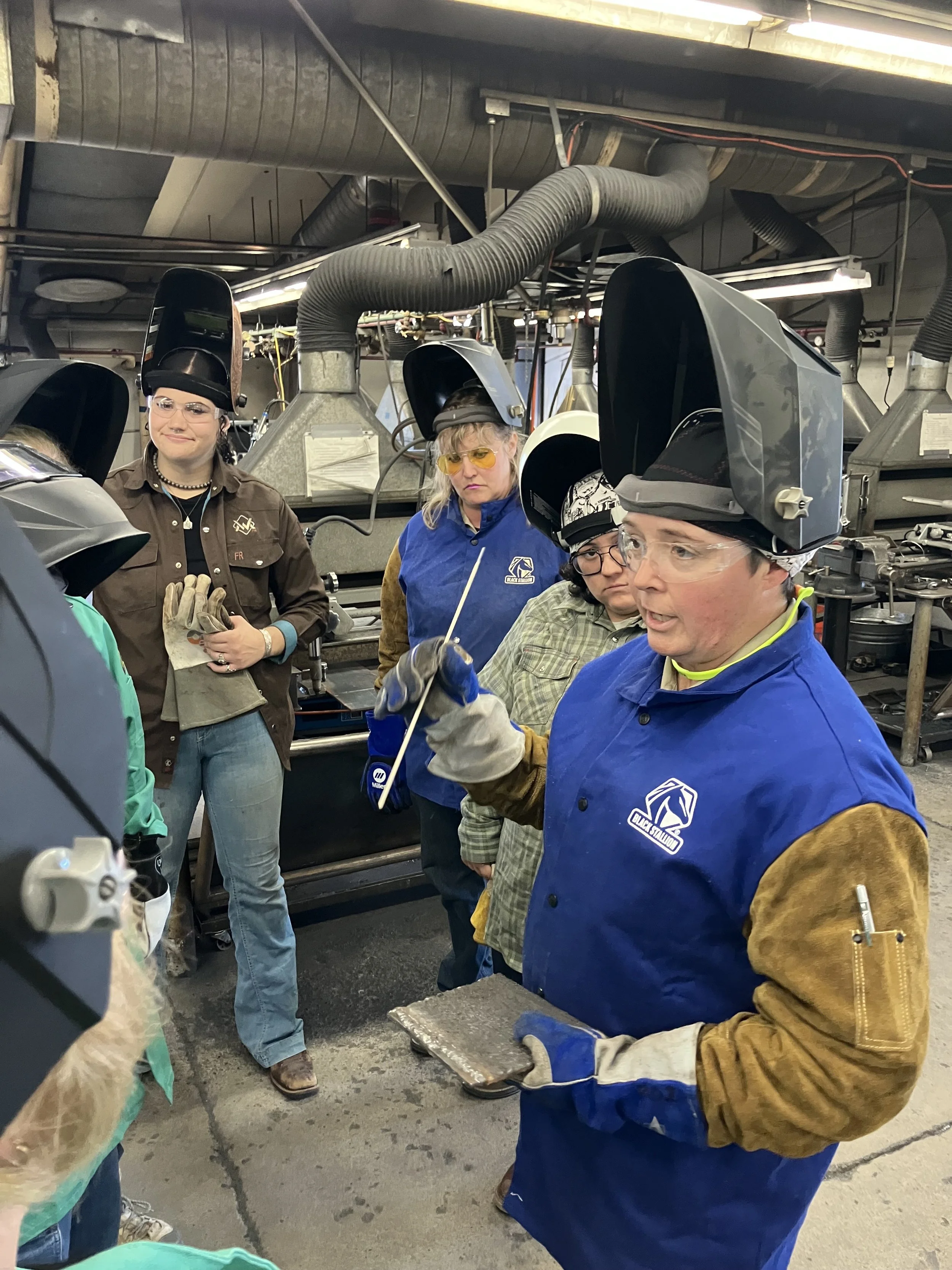 A group of women wearing welding protective gear in an industrial workshop, with ventilation ducts visible overhead.