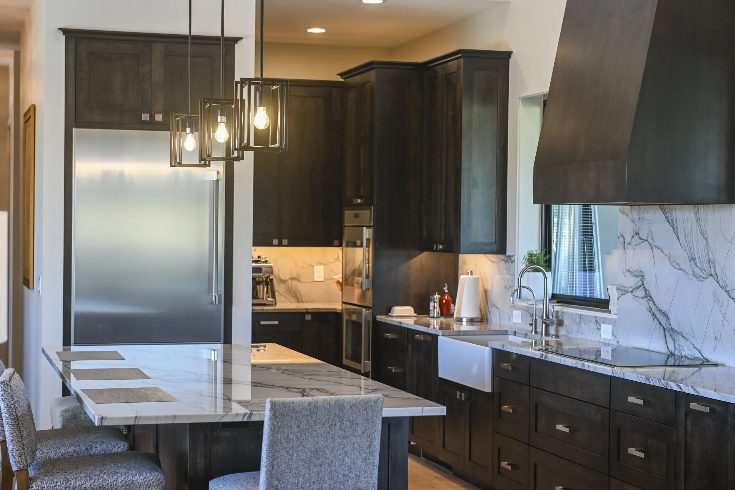 Modern kitchen with dark wood cabinets, marble island, stainless steel appliances, and a marble backsplash. There is a dining table with placemats and upholstered chairs, and a three-light pendant fixture hanging above.