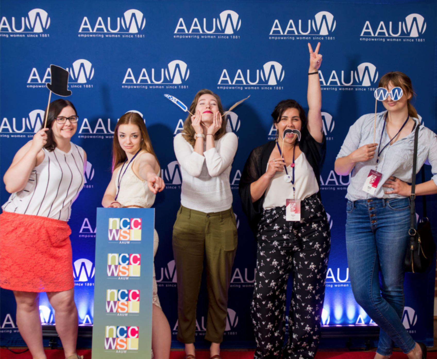Five women standing in front of a blue AAUW backdrop with logos that read 'empowering women since 1881.' They are holding various props including a top hat, glasses, a mustache, and a peace sign. The women are smiling and posing playfully, some pointing towards the camera.