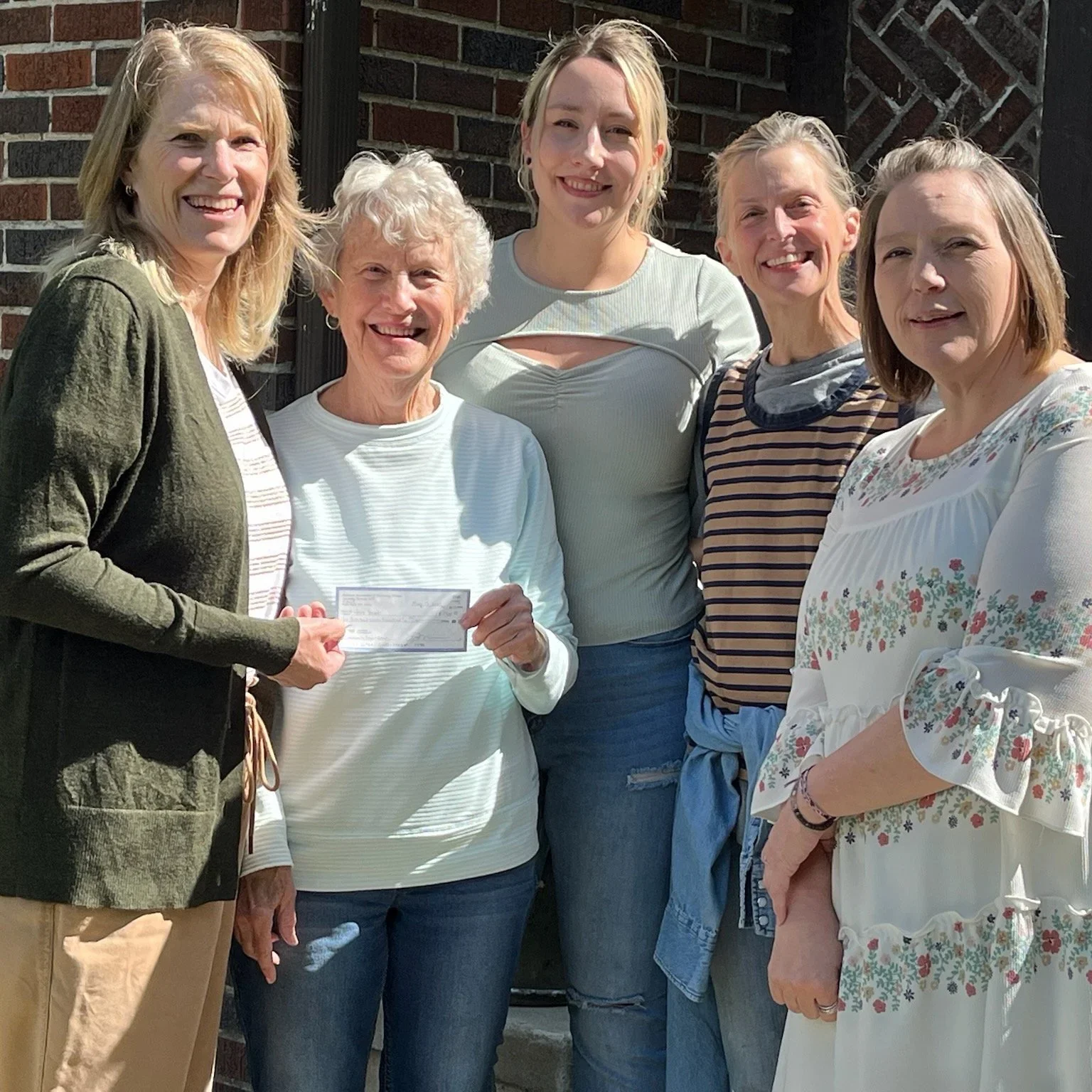 Group of six women standing outdoors, smiling, with brick wall background, one woman holding a check.