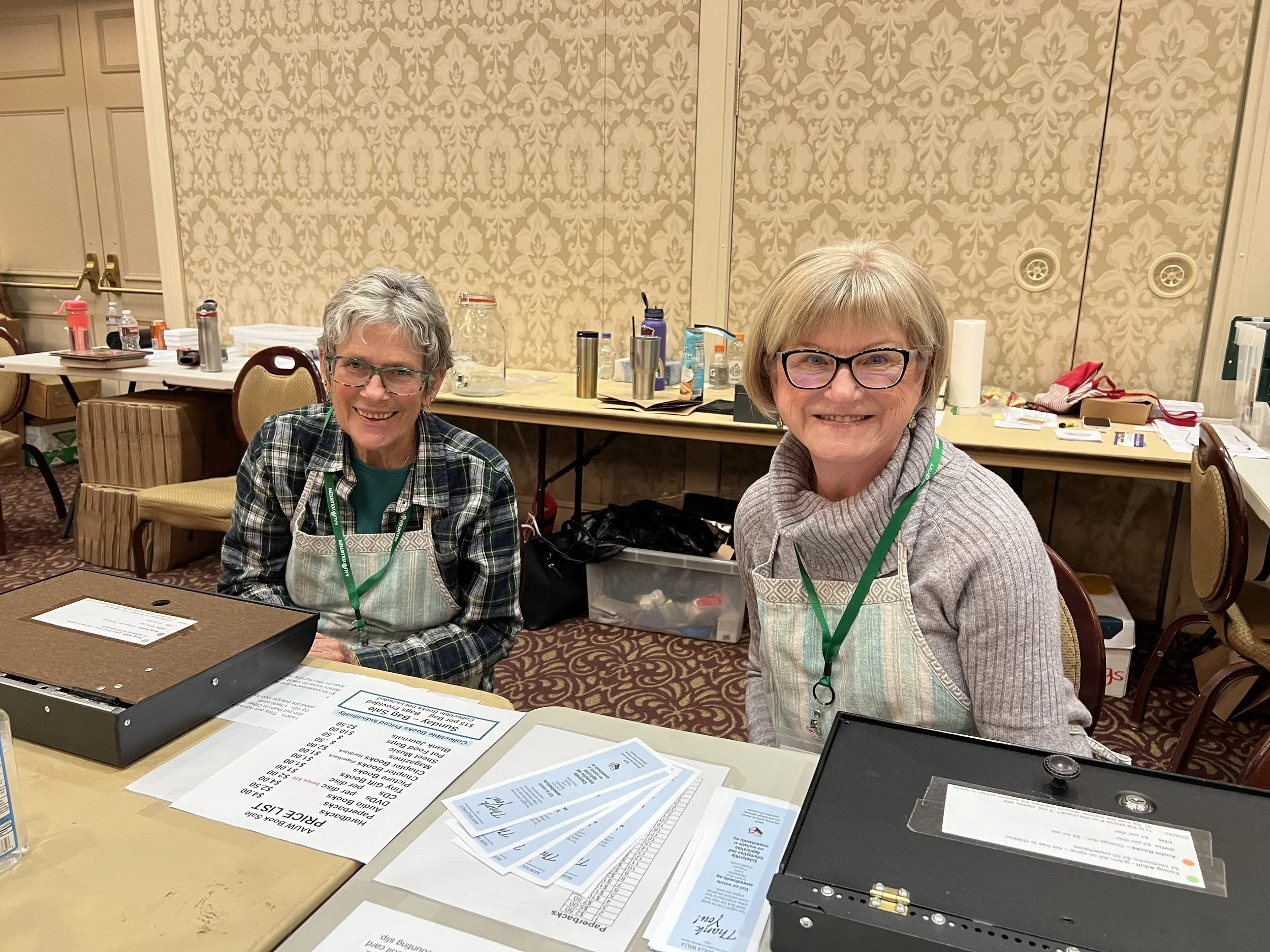 Two smiling women with short gray hair and glasses sitting at a table in a conference room, with paperwork, bottled water, and a black electronic device on the table, behind patterned wallpaper and a long table with more supplies.