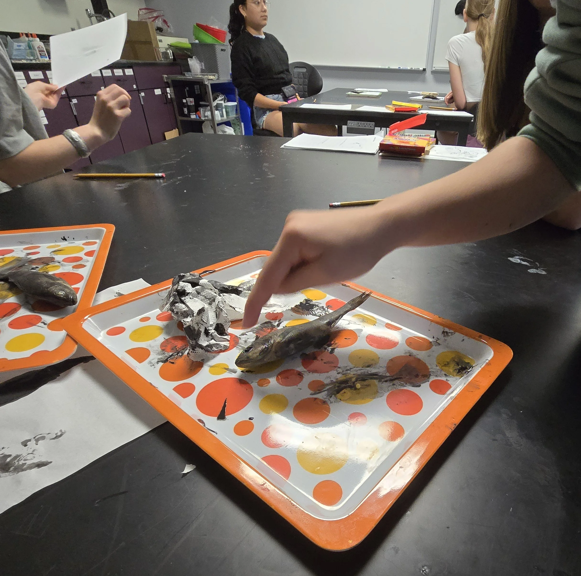 Students seated around a table in a classroom, with educational materials and a whiteboard in the background.