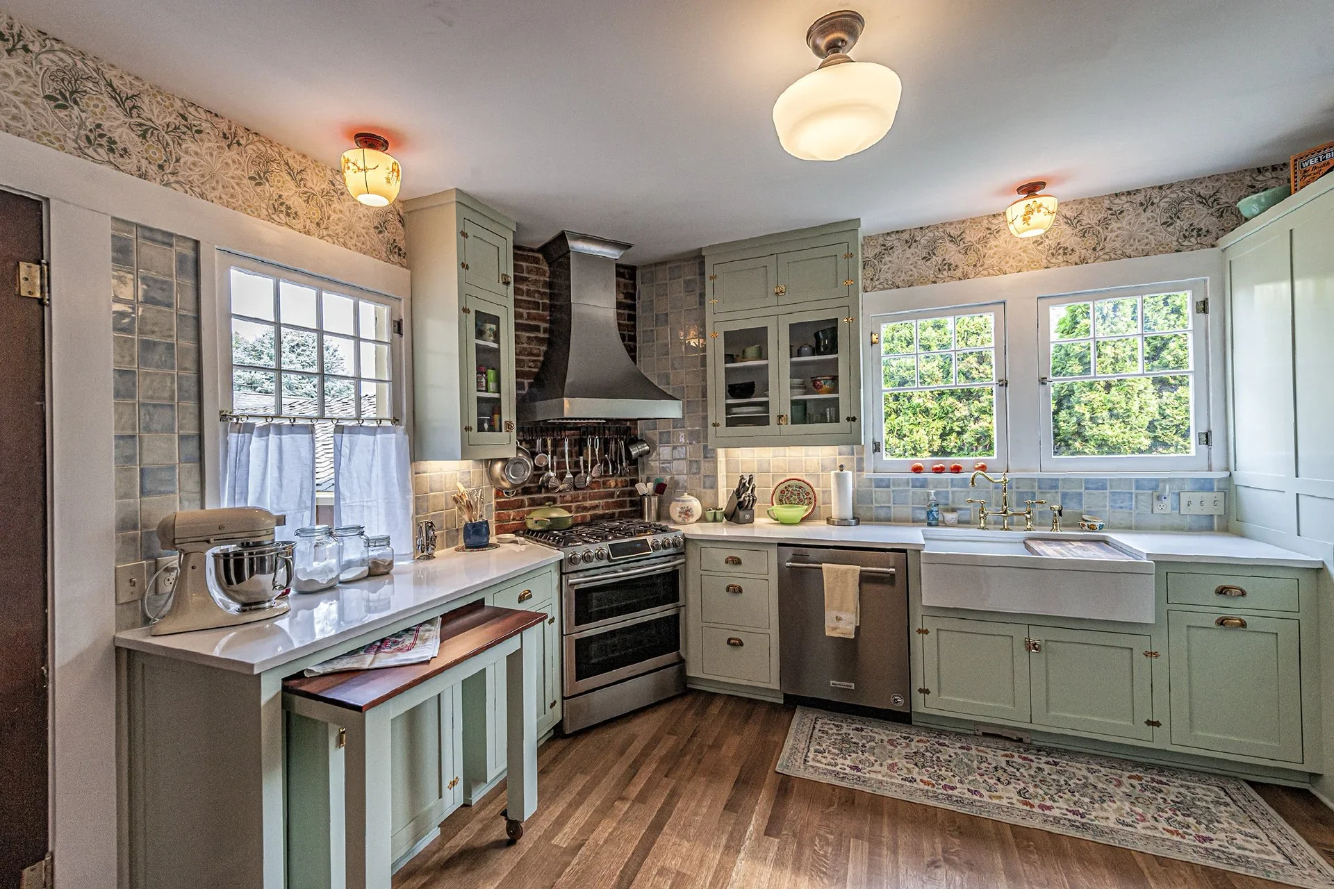 A vintage-style kitchen with mint green cabinets, a brick and tile backsplash, a stainless steel stove and range hood, and a window over the sink showing green trees outside.