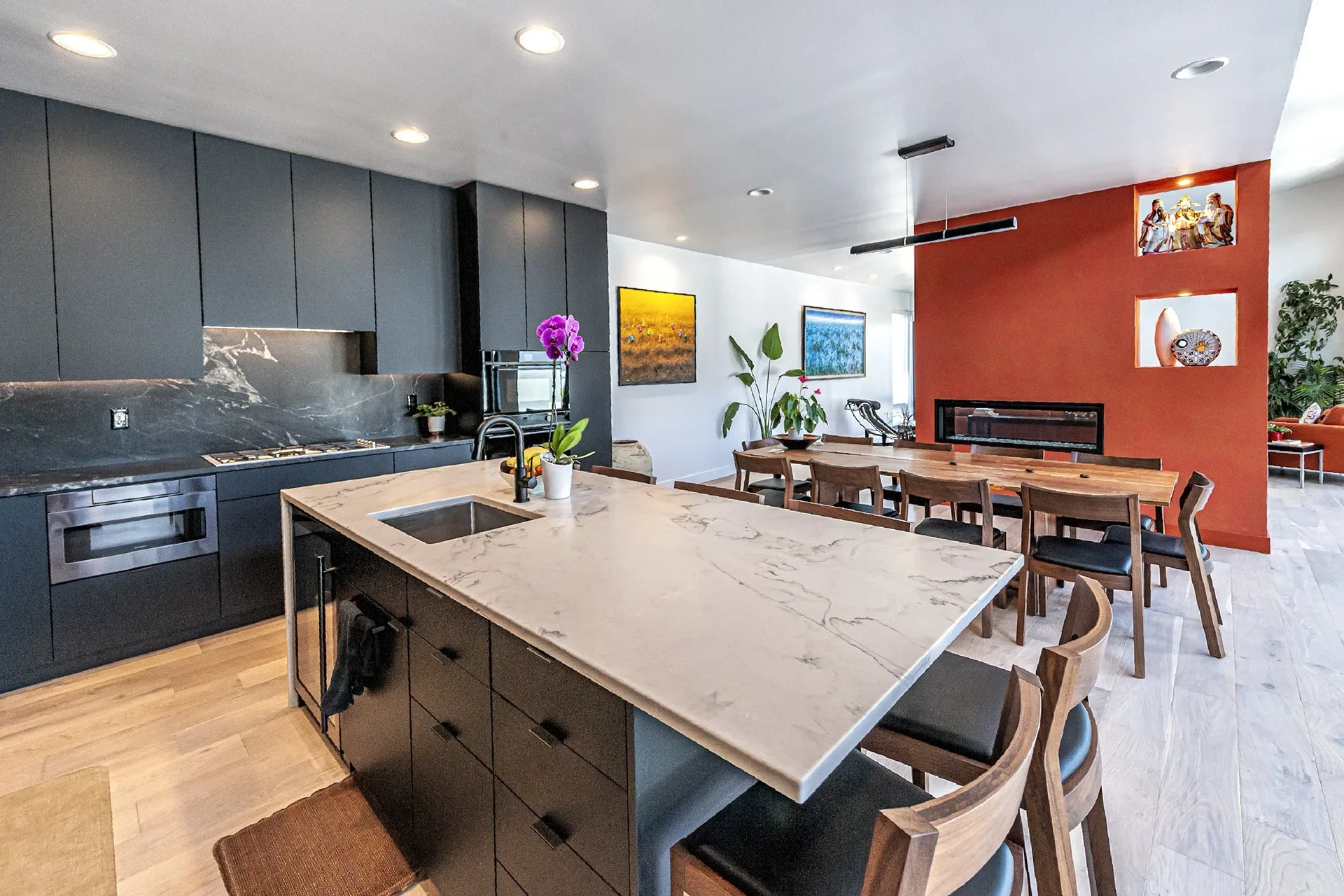 Open-concept kitchen and dining area with dark cabinets, marble countertops, a marble island, dining table with wooden chairs, colorful artwork on the walls, and decorative built-in niches with statues and a vase.