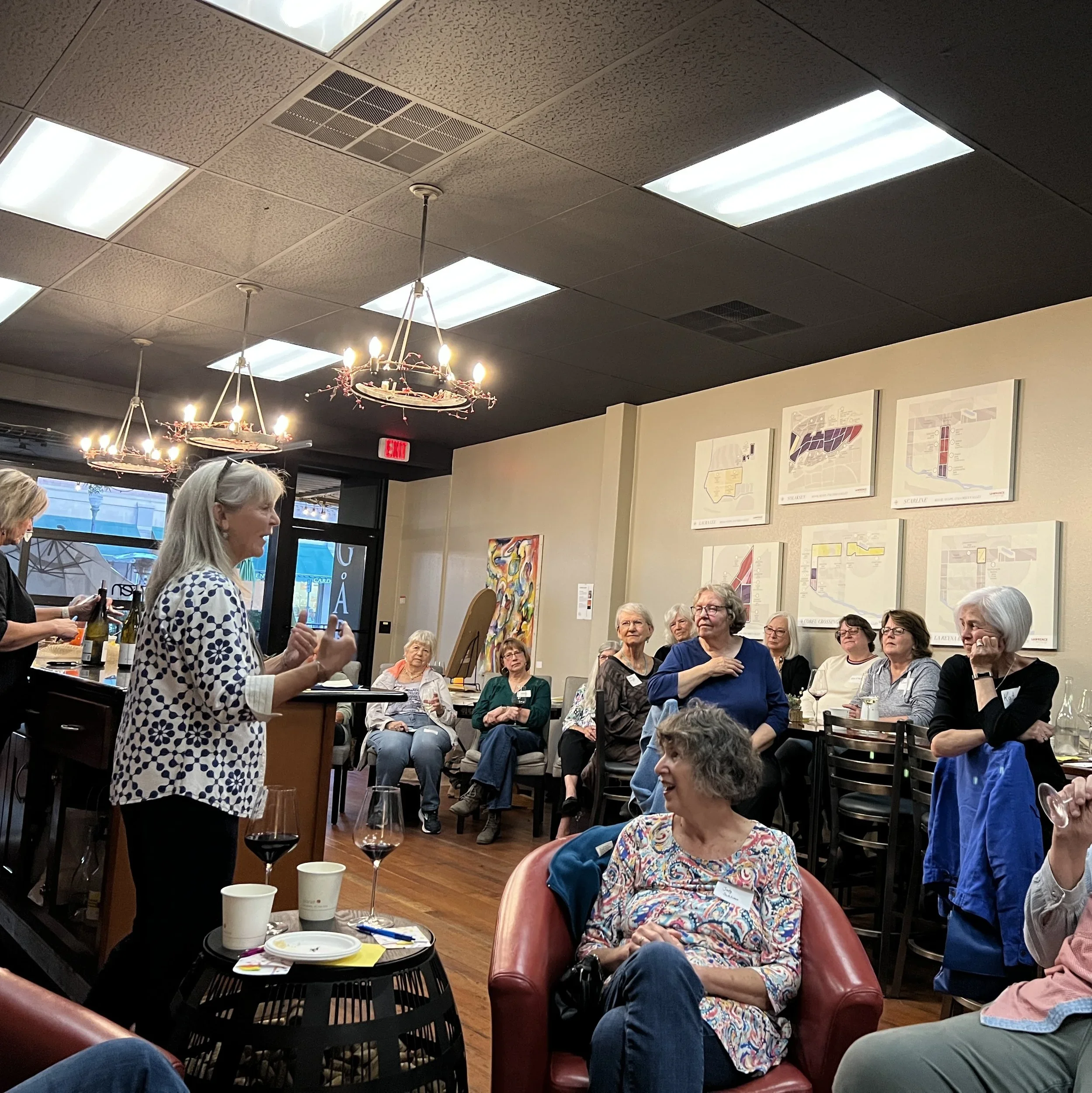 A group of people gathered, some seated around tables and others standing, engaged in conversation and listening. The room has framed art on the walls, bright ceiling lights, and decorative chandeliers.