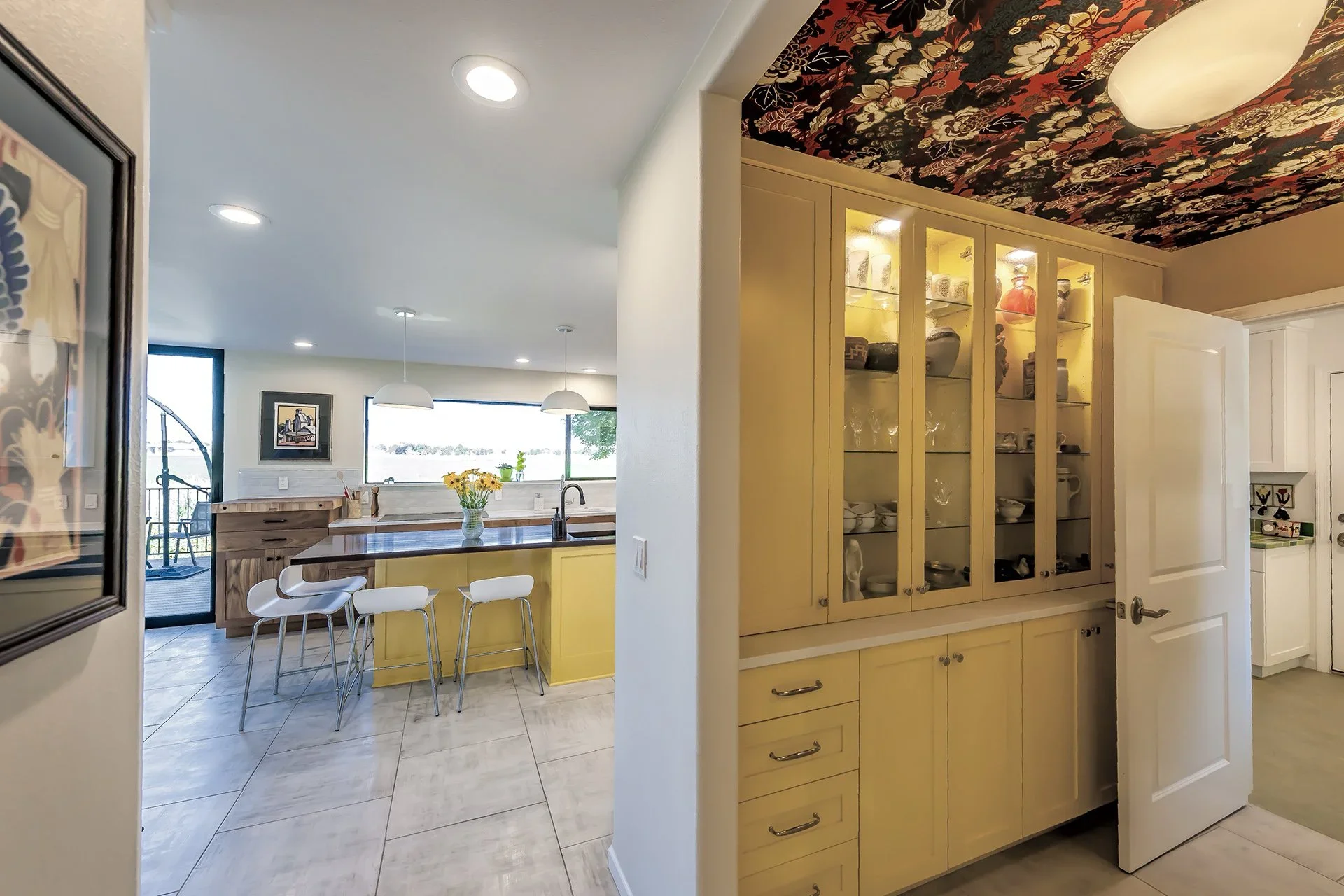 Kitchen with yellow cabinets, a wooden island with white bar stools, and a built-in glass-front cabinet displaying dishes, with tiled flooring and pendant lights over the island.
