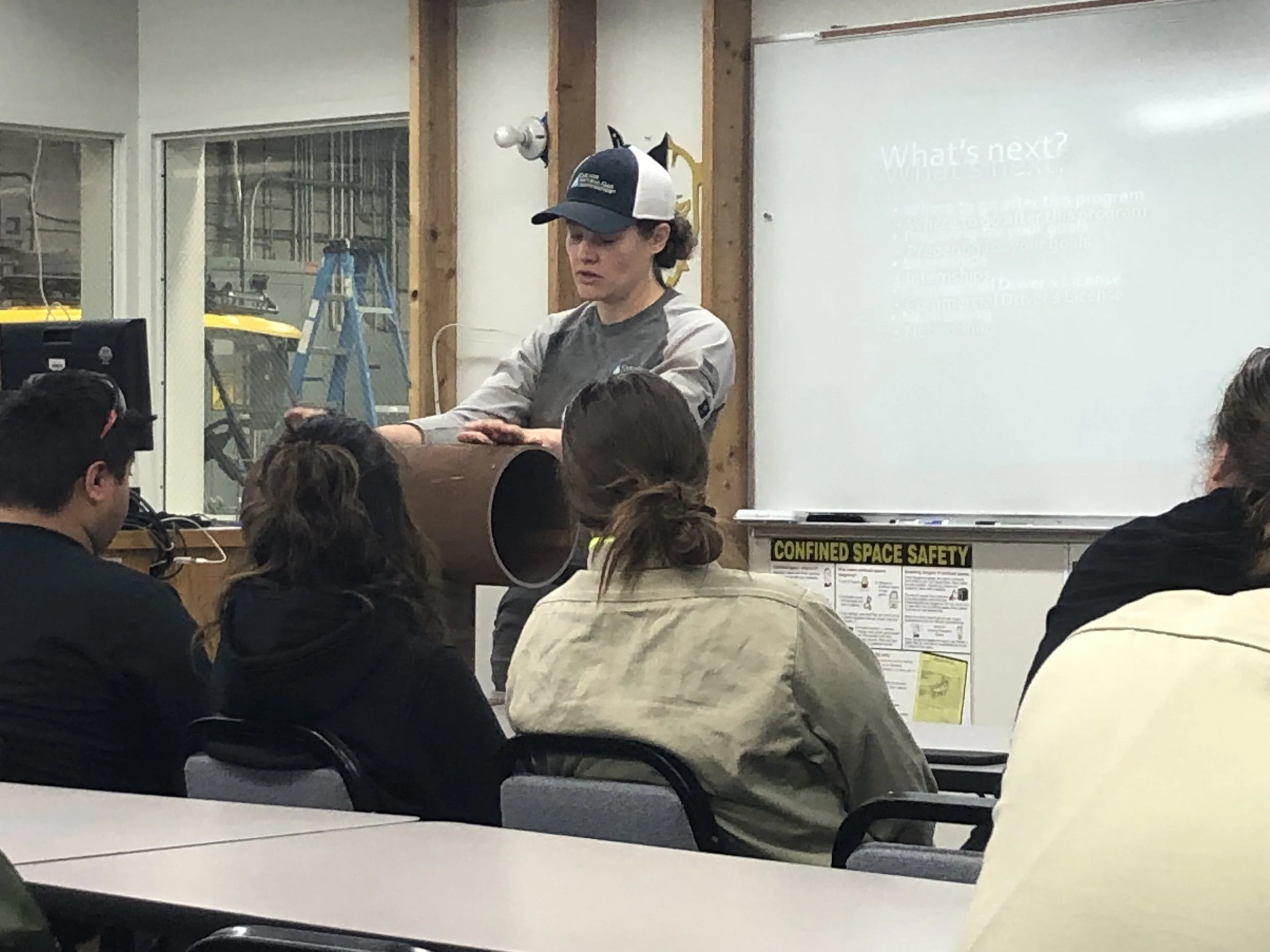 A person wearing a baseball cap stands in front of a whiteboard giving a presentation to a seated audience in a room with wooden beams and safety signs.